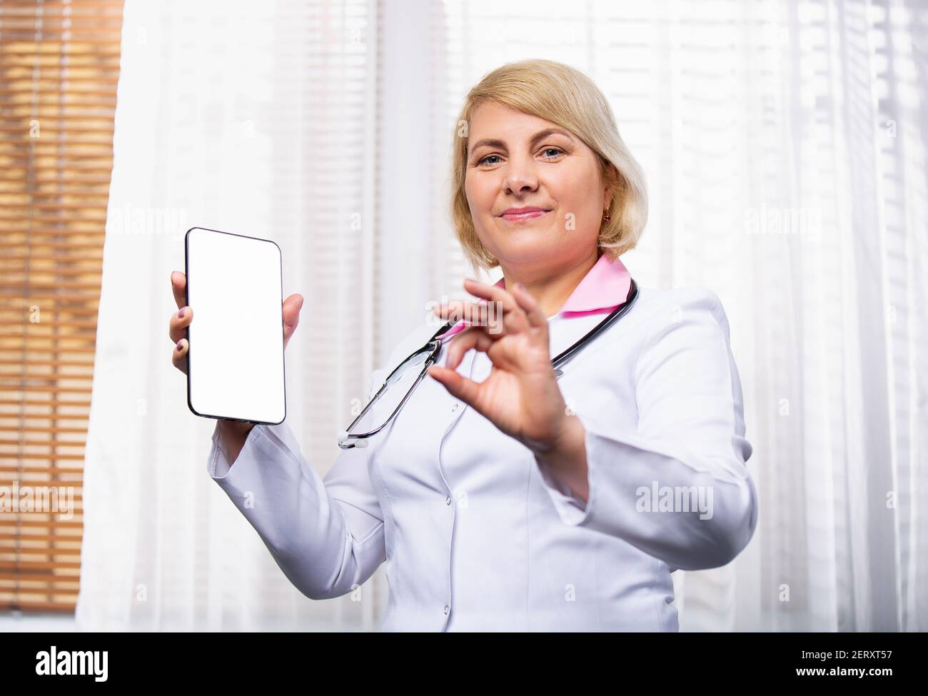 Medizinische Online-Beratung. Eine Frau in einem medizinischen Kleid zeigt einen weißen Bildschirm und zeigt ein Schild mit ihrer Hand, alles ist in Ordnung. Stockfoto