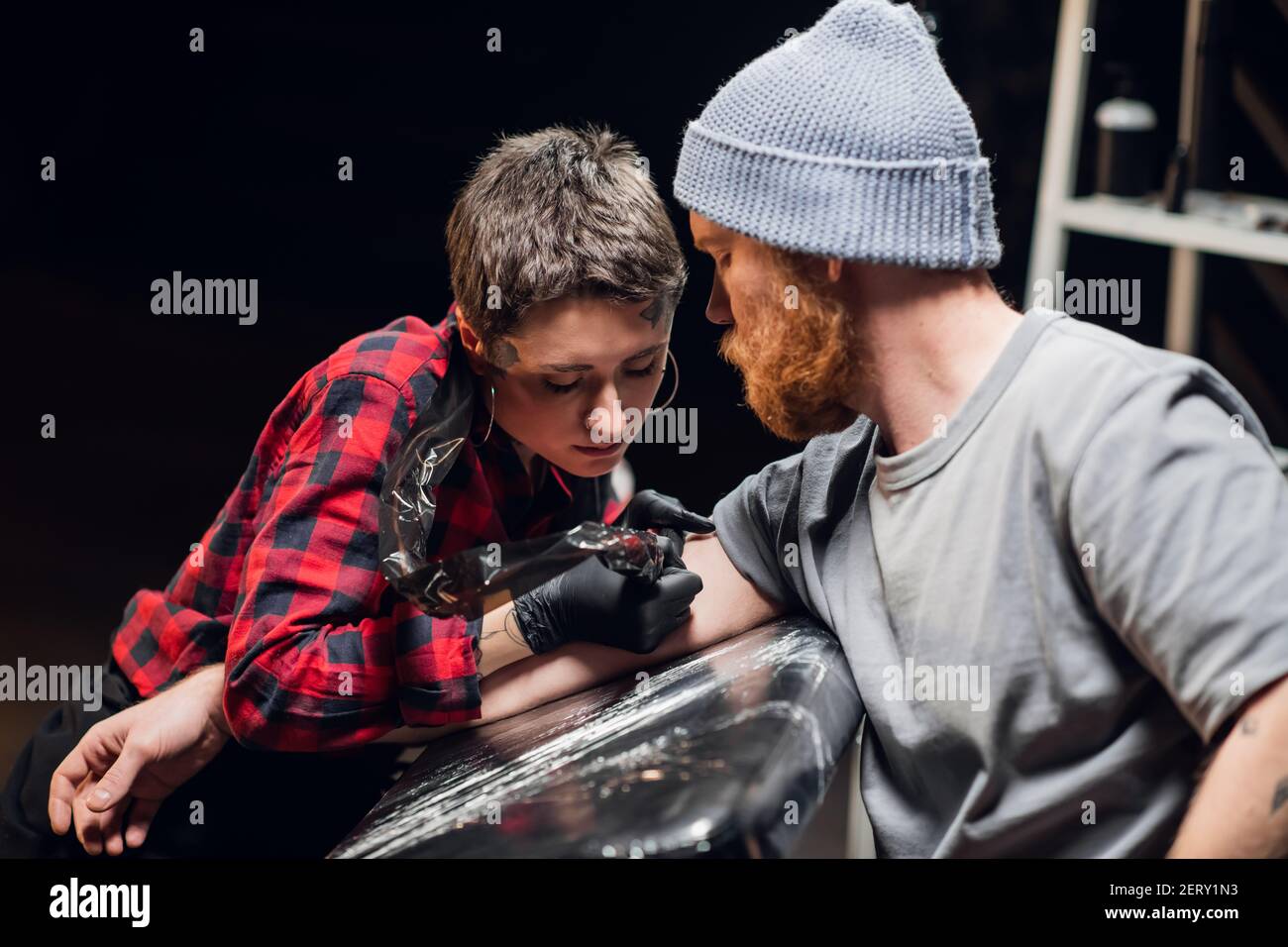 In einem modernen Tattoo-Salon macht ein Mädchen mit Piercings und Tattoos am Körper ein Tattoo auf dem Bizep einer jungen Hipster-Hand. Stockfoto