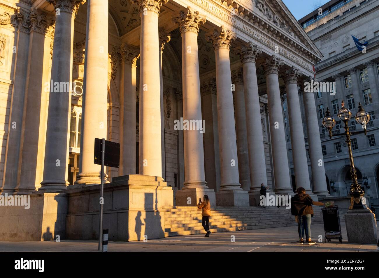 Während die meisten Londoner noch von zu Hause aus arbeiten, unterhalten sich Freunde unter den Säulen von Royal Exchange, während sie während der abendlichen Rush-Hour während der dritten Sperre des Coronavirus in der City of London, dem Finanzviertel der Hauptstadt, am 26th. Februar 2021 in London, England, entlang einer ruhigen Threadneedle Street spazieren. An der Spitze der dorischen und ionischen Säulen mit ihren verzierten Steinarbeiten kreuzen sich kraftvolle, starke Oberschwellen, die die Last feiner Kunst und Schnitzereien tragen, die das Design von Sir William Tite im Jahr 1842-1844 zeigen und 1844 von Königin Victoria eröffnet wurden, deren Name in lateinischer Sprache (Victoria Stockfoto