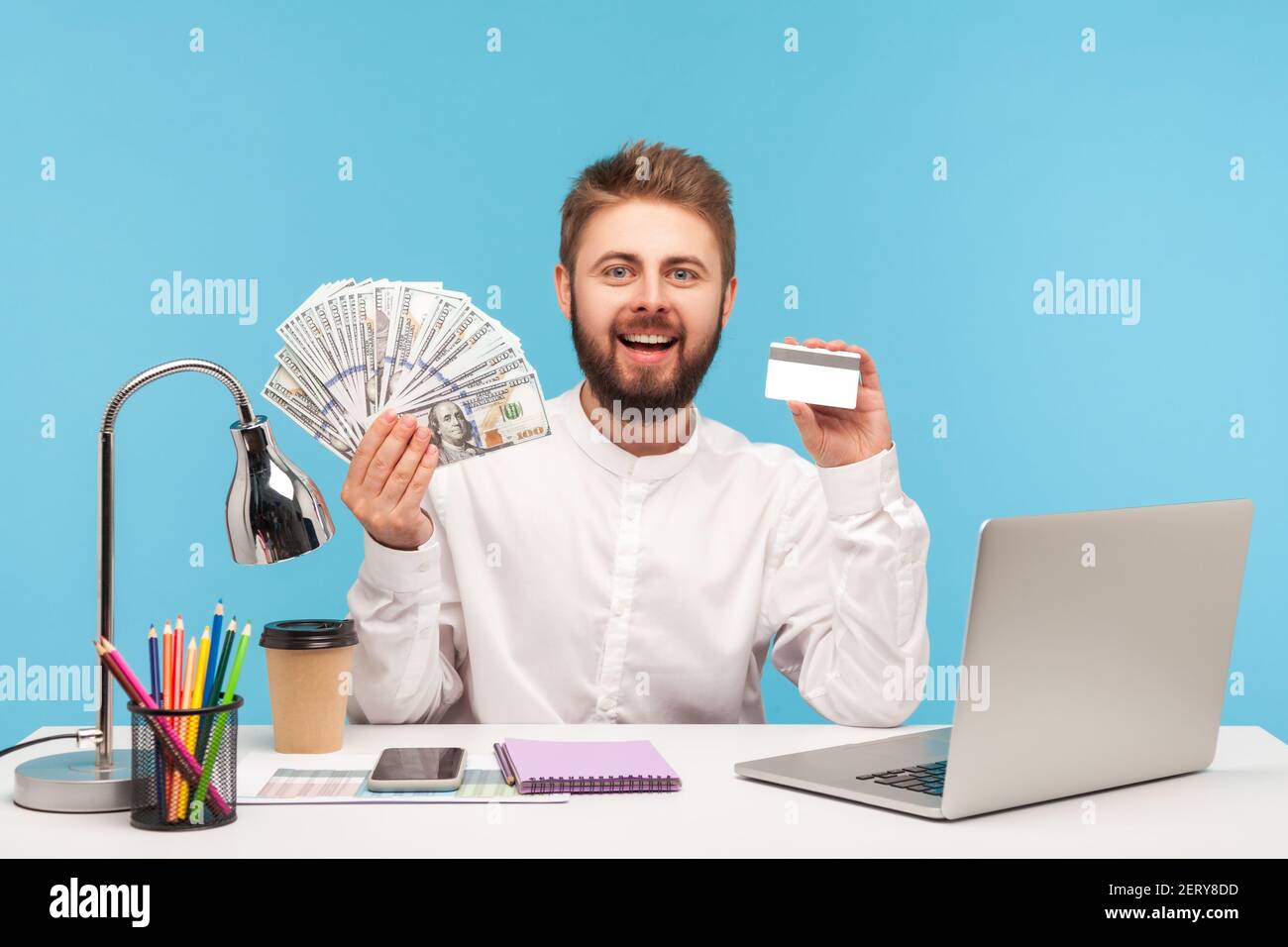Glücklicher lächelnder Mann Büroangestellter hält und zeigt Dollar Bargeld und Kreditkarte sitzen am Arbeitsplatz mit Laptop, Banküberweisung, Gutschrift. Innen Stockfoto