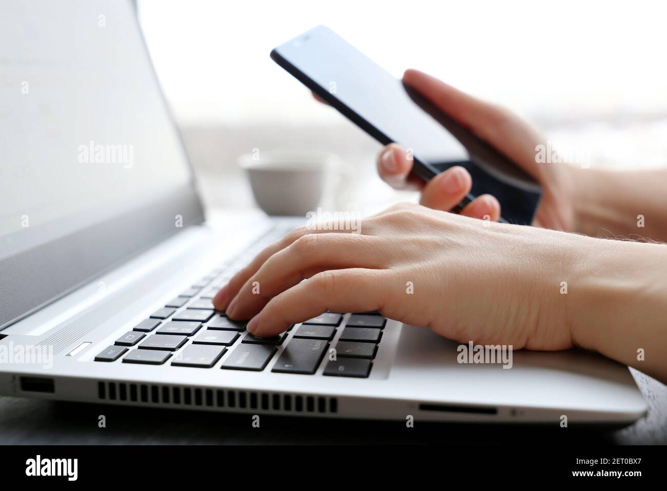 Frau mit Smartphone sitzt auf Laptop-Tastatur in der Nähe des Fensters. Konzept der Online-Kommunikation, Büro- oder Heimarbeit und Bezahlung Stockfoto