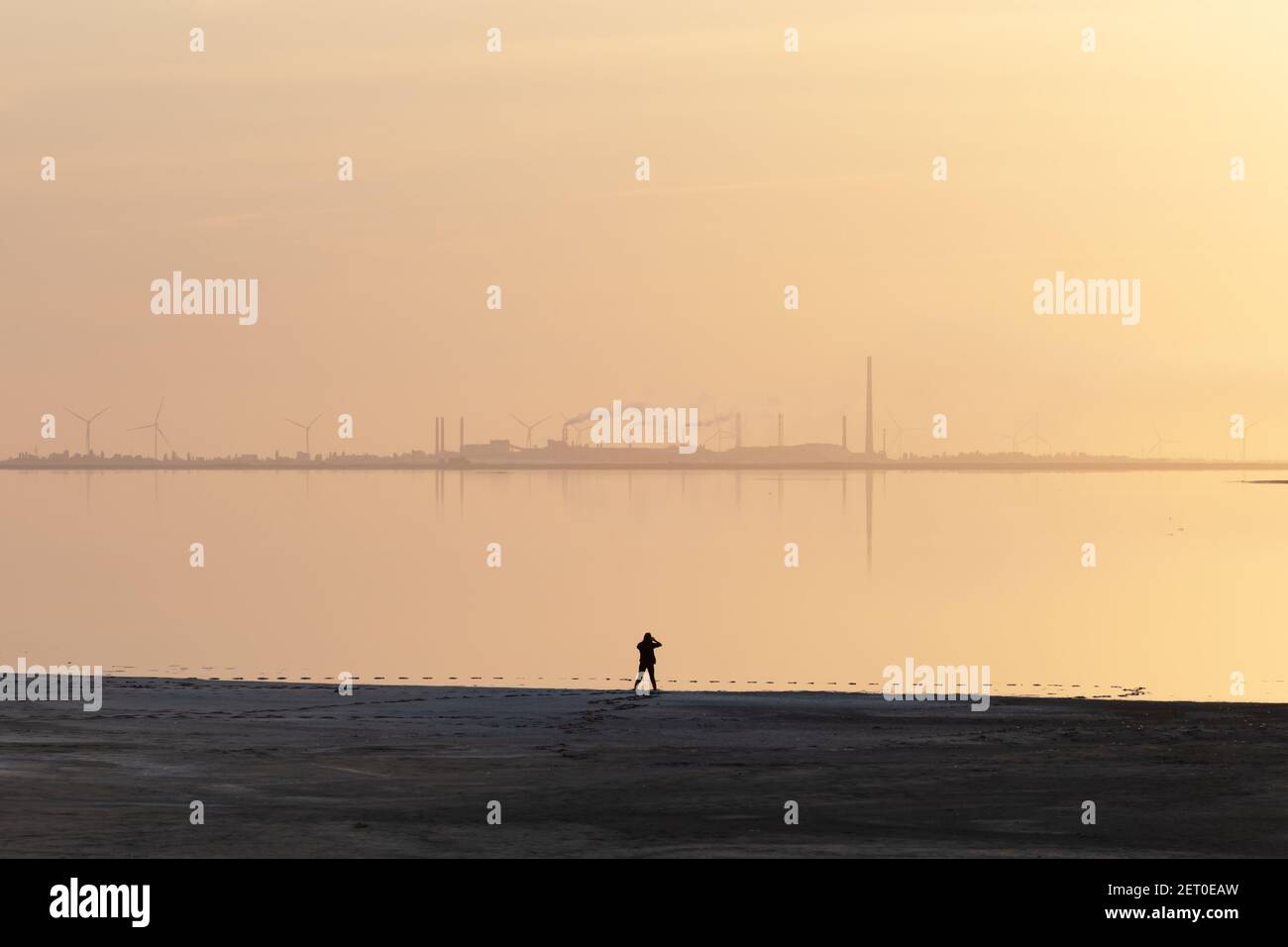 Ruhige minimalistische Landschaft mit einsamen Mädchen auf ruhigem Wasser und Industriegebiet mit Fabriken und Windturbinen im Hintergrund. Minimale Landschaftsfotografie Stockfoto