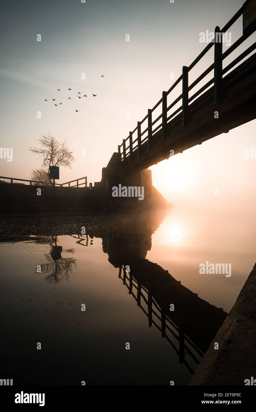Sonnenaufgang von unter Holzfußbrücke über Fluss reflektiert Im Kanal schöne orange Reflexion Kristall noch ruhige Wasservögel Fliegen in den Himmel Stockfoto