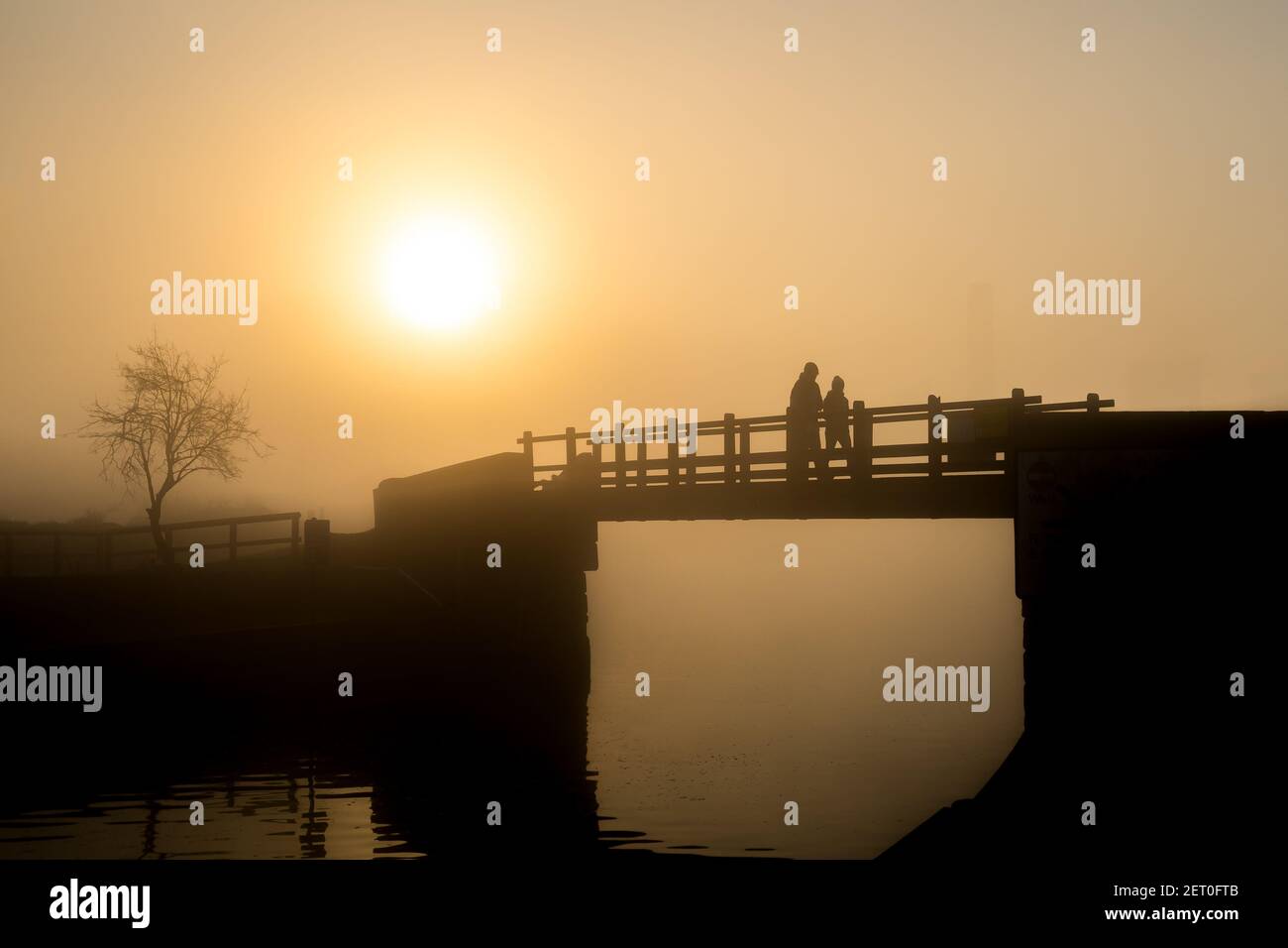 Erstaunlich hell orange Nebel Sonnenaufgang in Liebe Paar Silhouette suchen Bei Blick im Nebel Achtsamkeit ruhige hölzerne Fußbrücke über Friedlicher Flusskanal Stockfoto