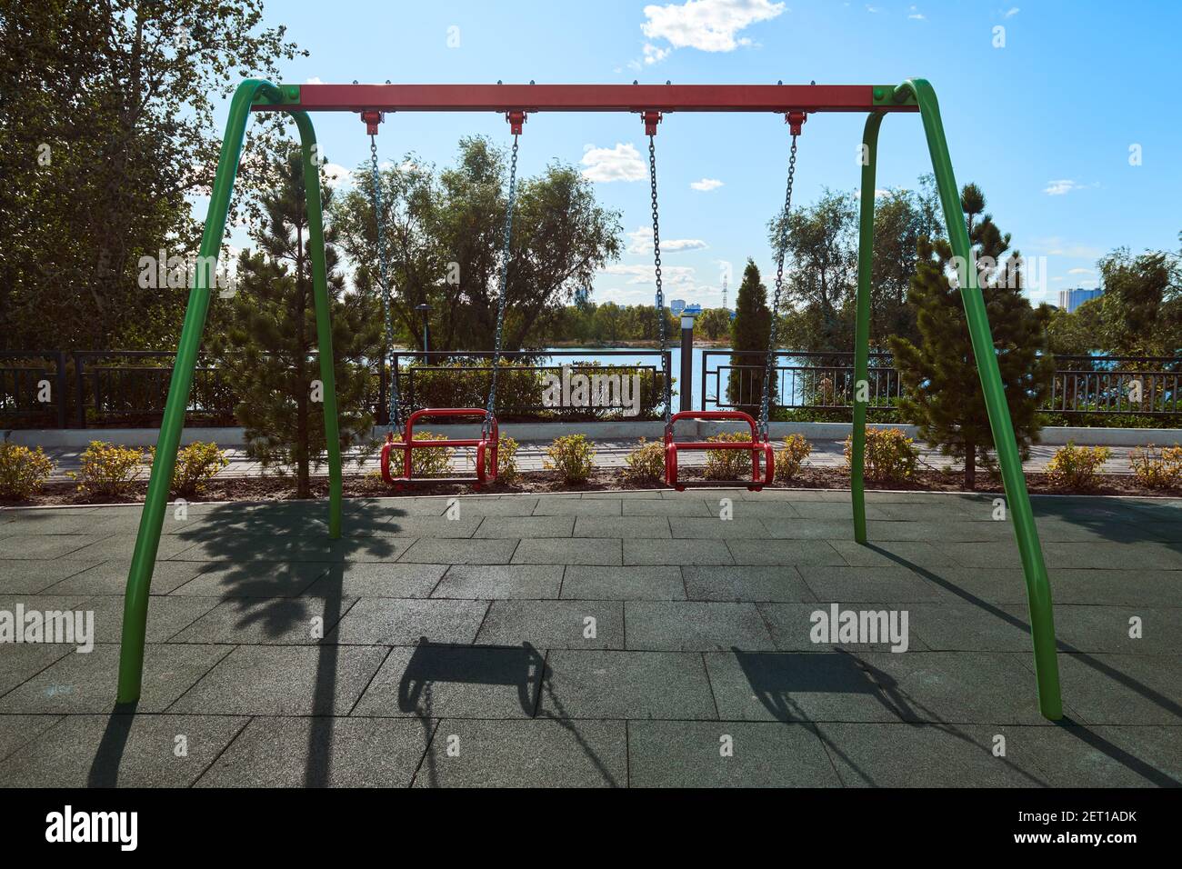 Leere rote Schaukel auf dem Spielplatz gegen blauen Himmel und Bäume. Schaukel hängt an den Metallketten. Leerer Platz während der Coronavirus-Pandemie. Covid-19. Stockfoto