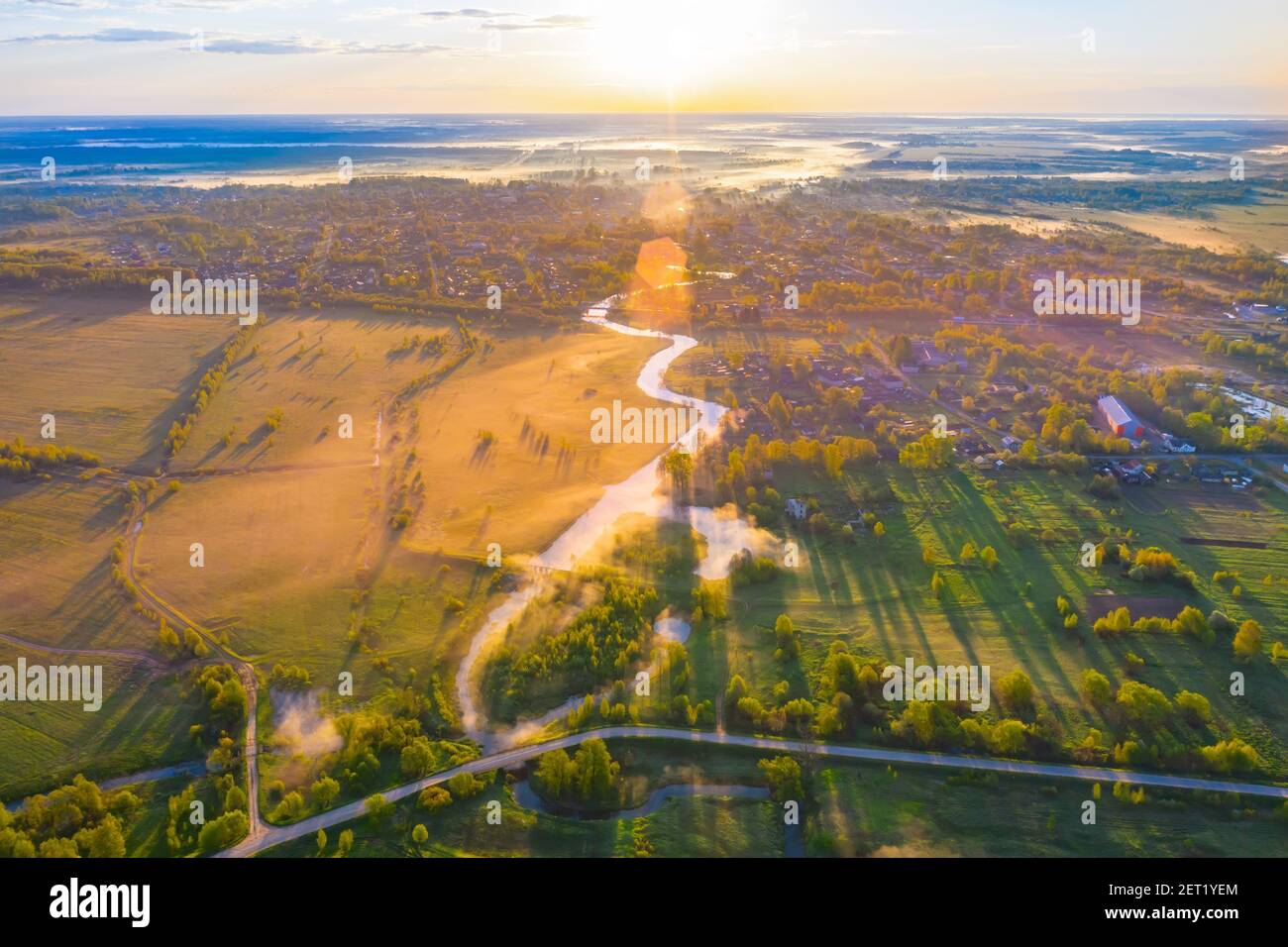 Fliegen über ein nebeliges Flusstal, nasse Wiesen und hohe Bäume, die einen Schatten von der Morgensonne werfen. Flug über eine kleine ländliche Stadt Stockfoto