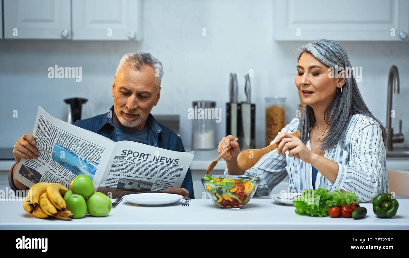 Ältere asiatische Frau Mischung Salat in der Nähe Ehemann Lesen Sport-Nachrichten Während des Frühstücks Stockfoto
