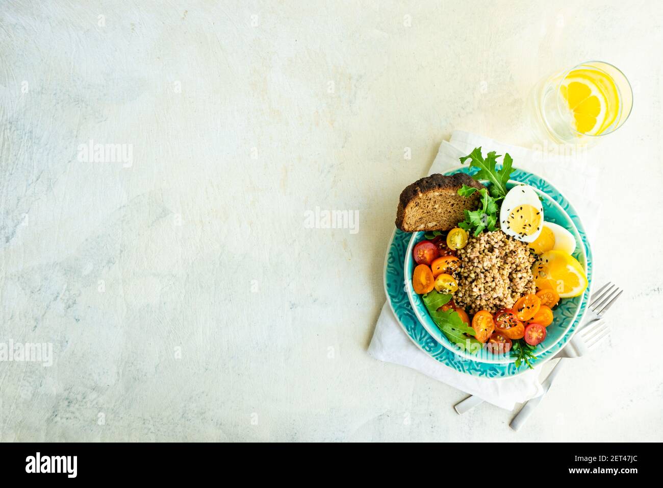Schüssel Buchweizensalat mit Rucola, Tomate, Salat, hart gekochtem Ei und einer Scheibe Brot neben einem Glas Zitronenwasser Stockfoto