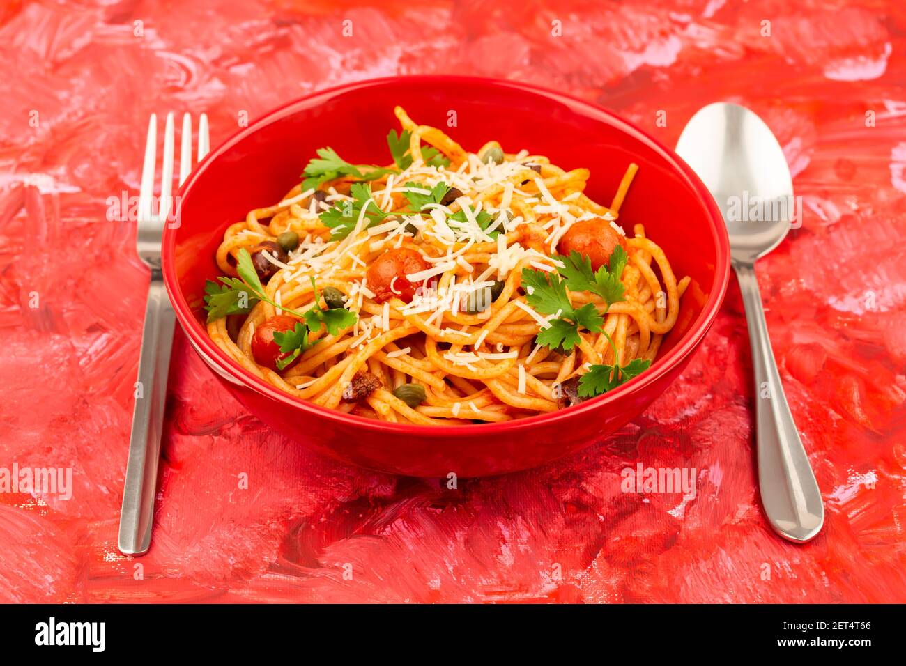 Pasta Spaghetti Puttanesca in roter Schale mit Gabel und Löffel auf buntem Hintergrund. Italienische Küche. Stockfoto