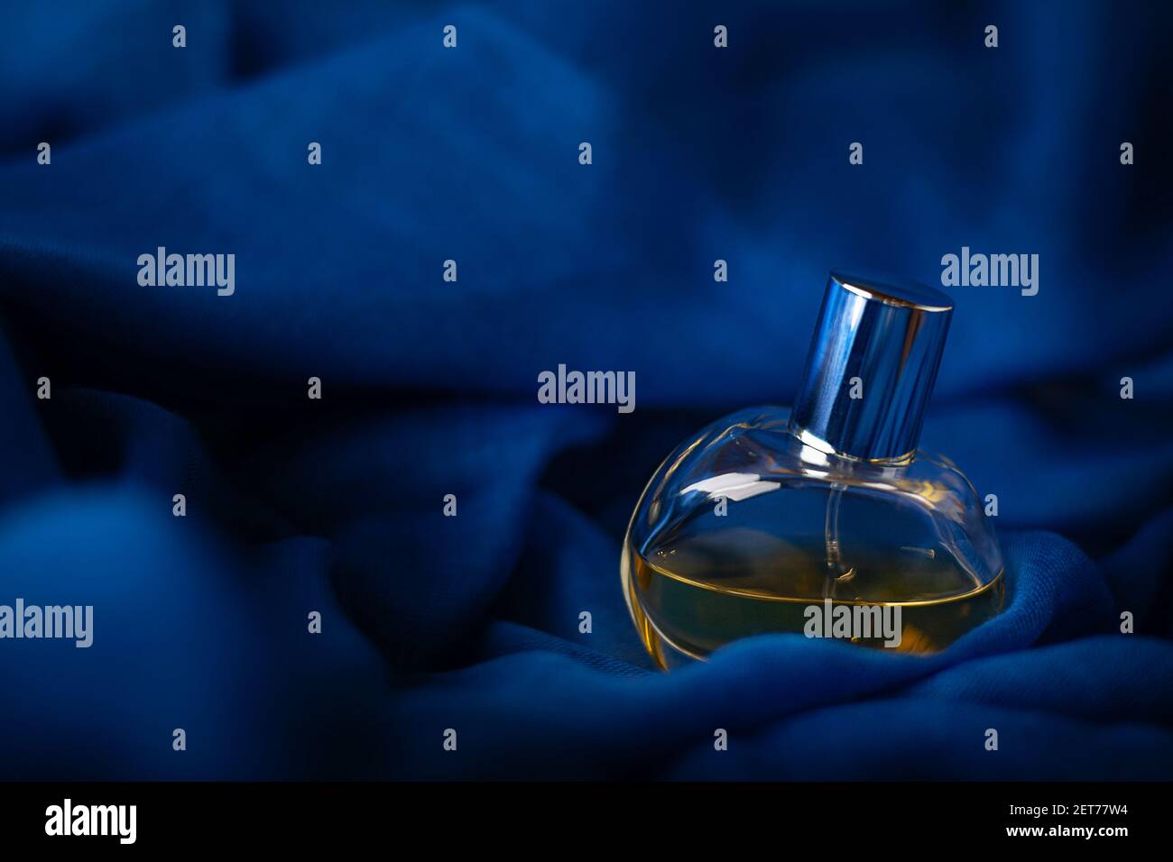 Transparente Glasflasche mit Parfüm auf blauem Stoff mit weichem Fokus. Nahaufnahme in dunklem Studio mit Kopierbereich. Das Konzept ist wunderschön, nachts Stockfoto