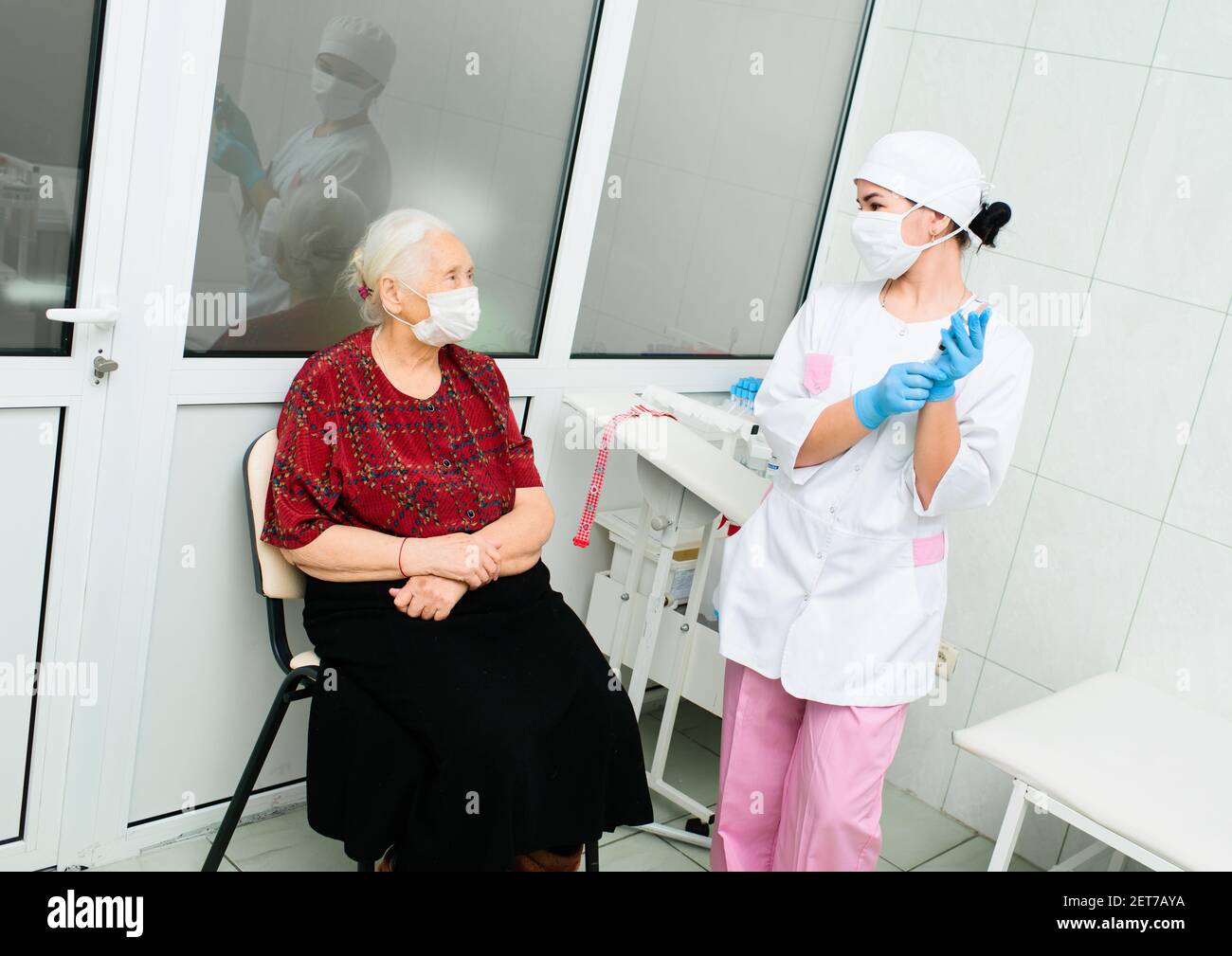 Krankenschwester in einer medizinischen Maske zieht Gummihandschuhe an und Bereitet sich auf die ärztliche Untersuchung des Patienten - und vor Ältere grauhaarige Frau Stockfoto