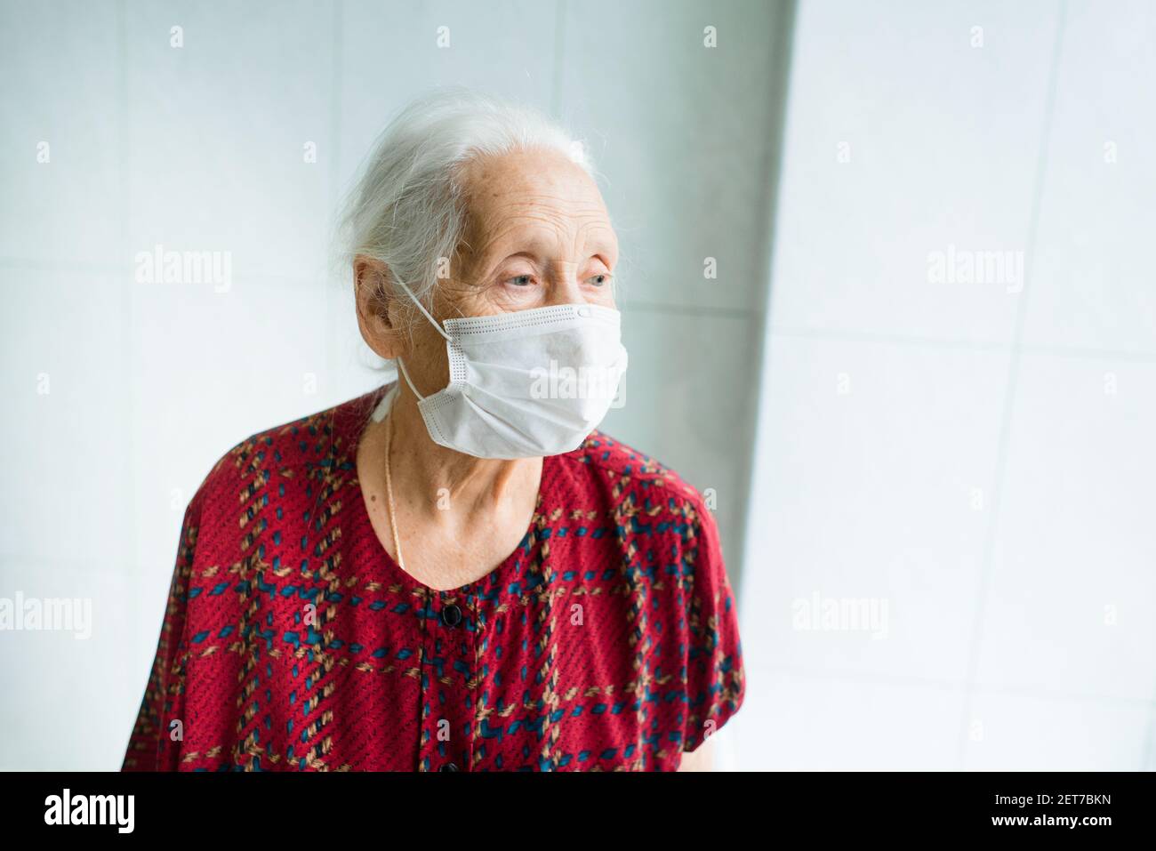 Eine ältere Frau mit grauem Haar 70-80 Jahre alt in einer medizinischen Maske allein im Zimmer steht am Fenster. Stockfoto Eine ältere Frau mit grauem Haar 70-80 Jahre alt in einer medizinischen Maske allein im Zimmer steht am Fenster. Stockfoto