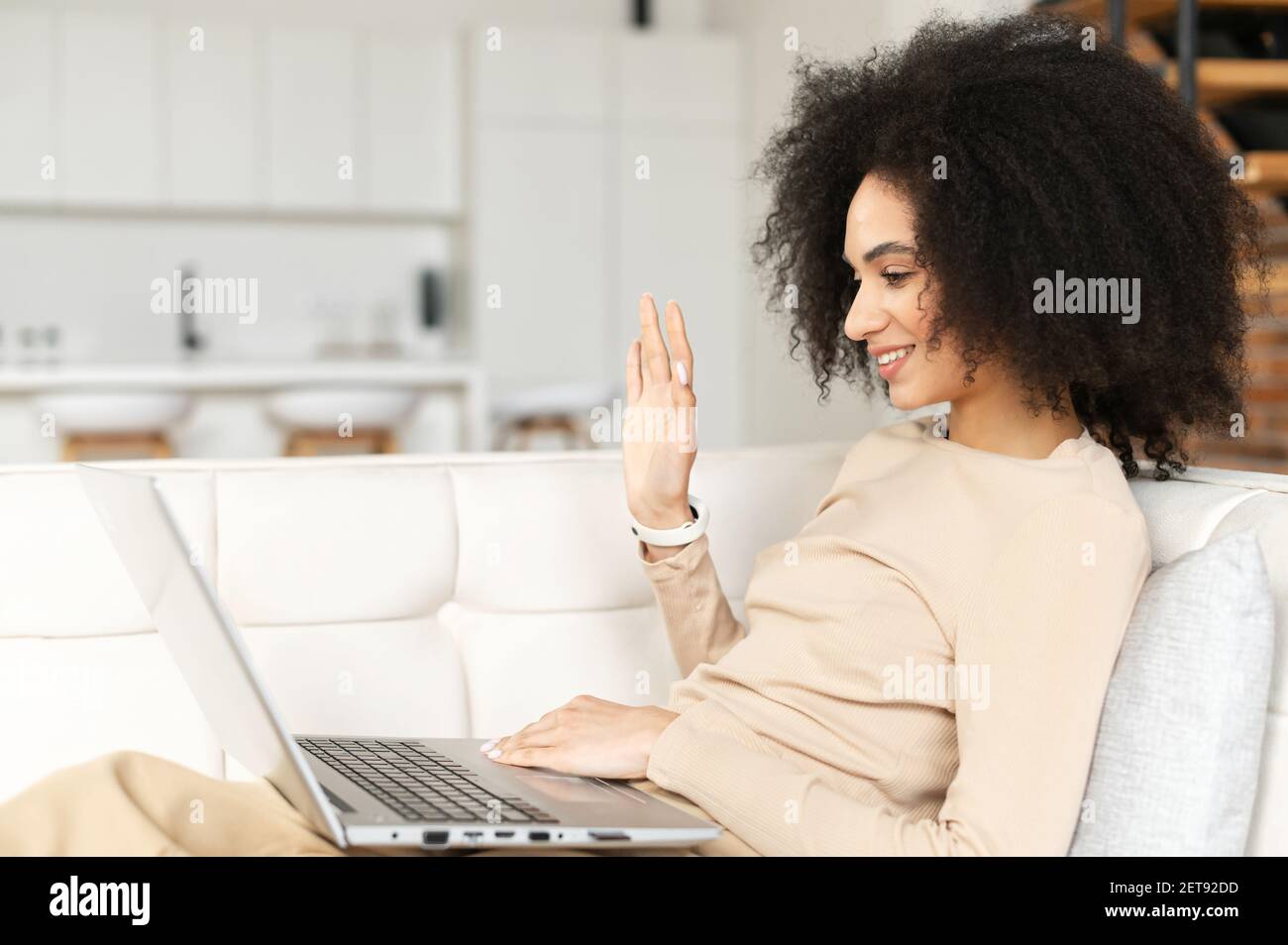 Videoanruf-Konzept. Fröhliche afroamerikanische Bloggerin mit voluminösen dunklen Haaren, winkende Hand, während sie von zu Hause aus einen Videoanruf auf dem Laptop hat, auf dem Sofa liegt, freiberuflich arbeiten, jederzeit und überall Stockfoto