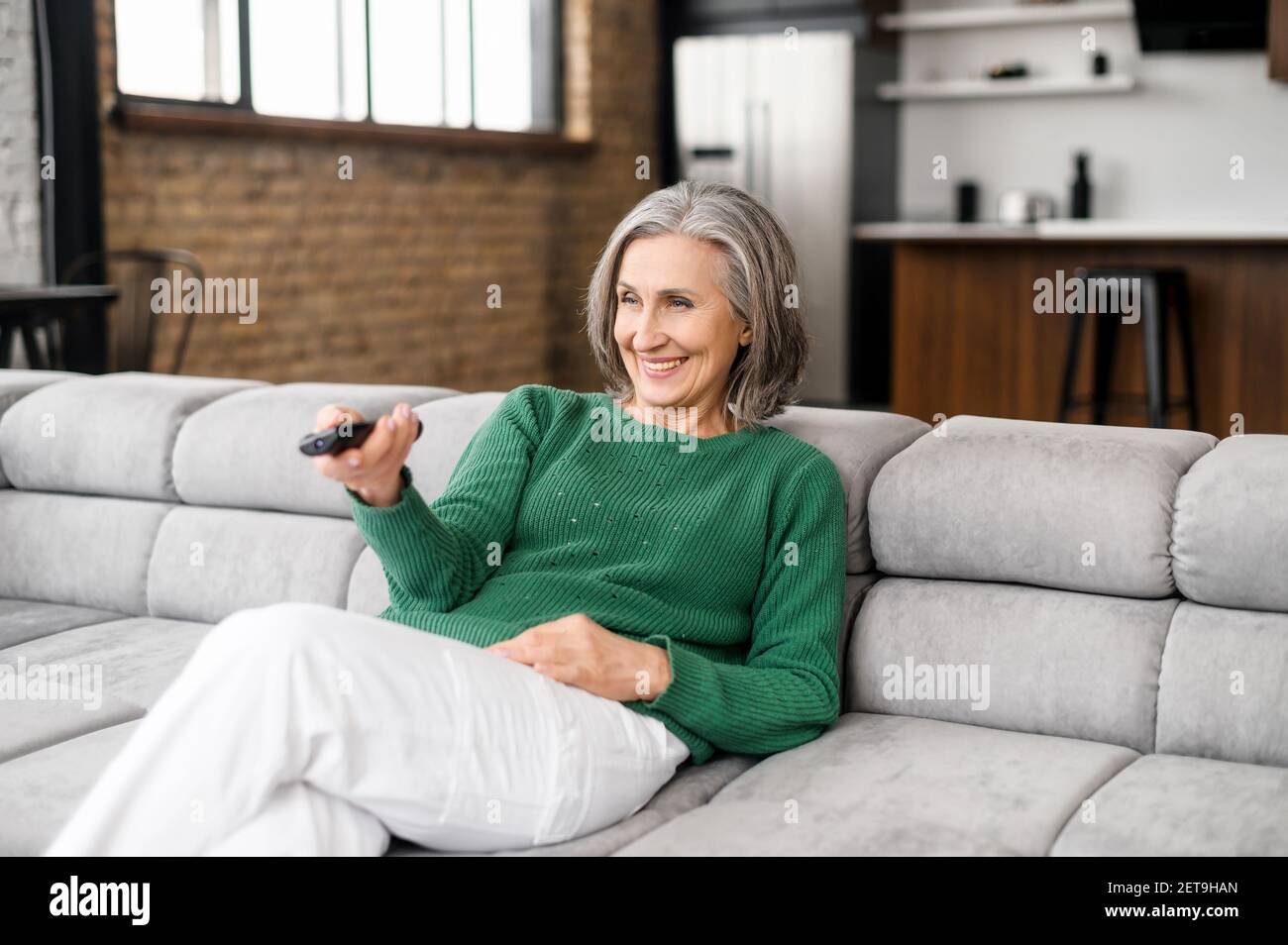 Glückliche lächelnde ältere Dame mit grauen Haaren in einem grünen Pullover sitzt auf der Couch im Wohnzimmer, zeigt eine tv-Fernbedienung, um die Kanäle zu wechseln, aufgeregt, um Lieblings-Film Online-Serie zu sehen Stockfoto