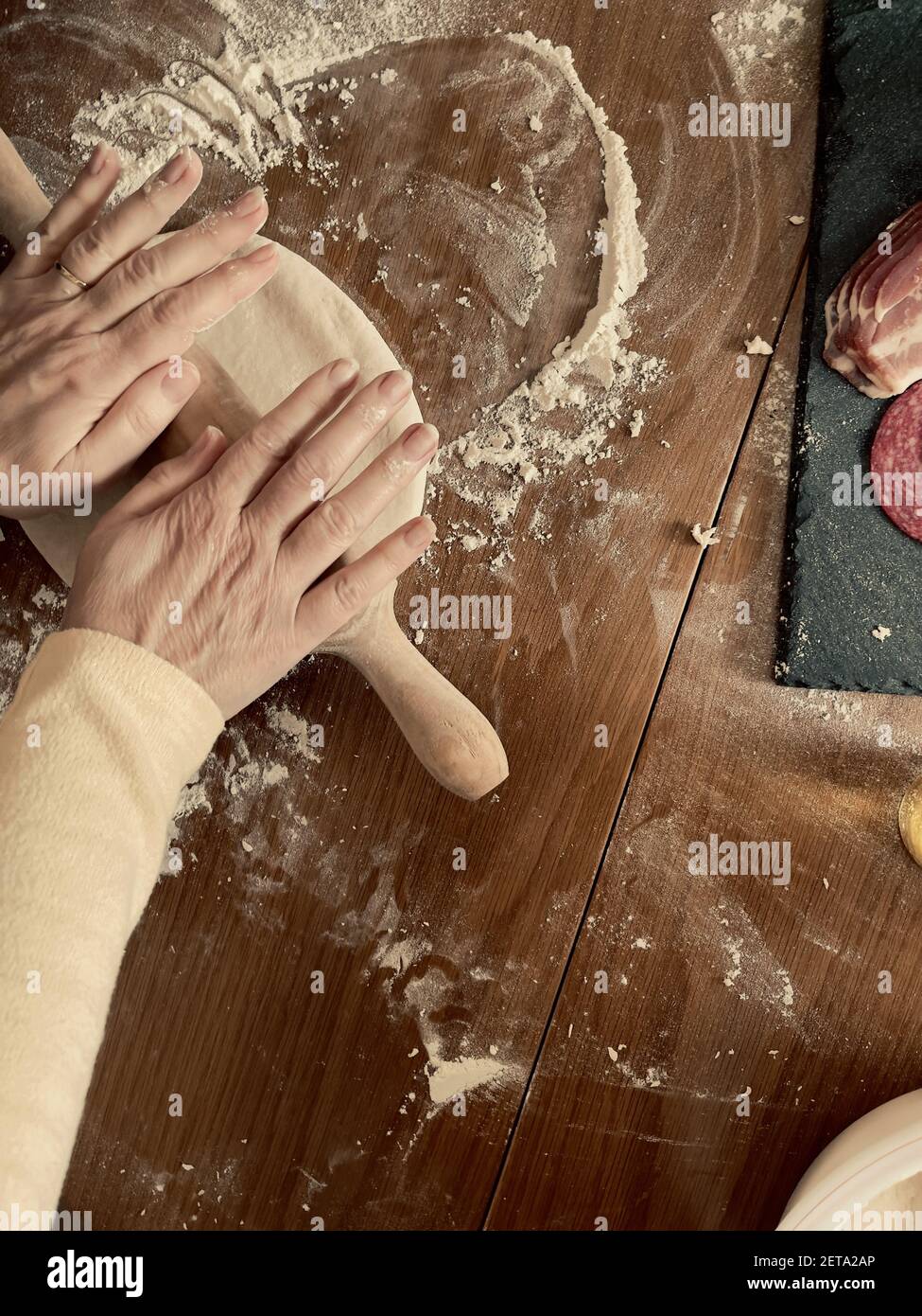 Eine alte Frau, die den Teig mit einem Holzrollen ausdehnt PIN auf dem mehligen Tisch Stockfoto