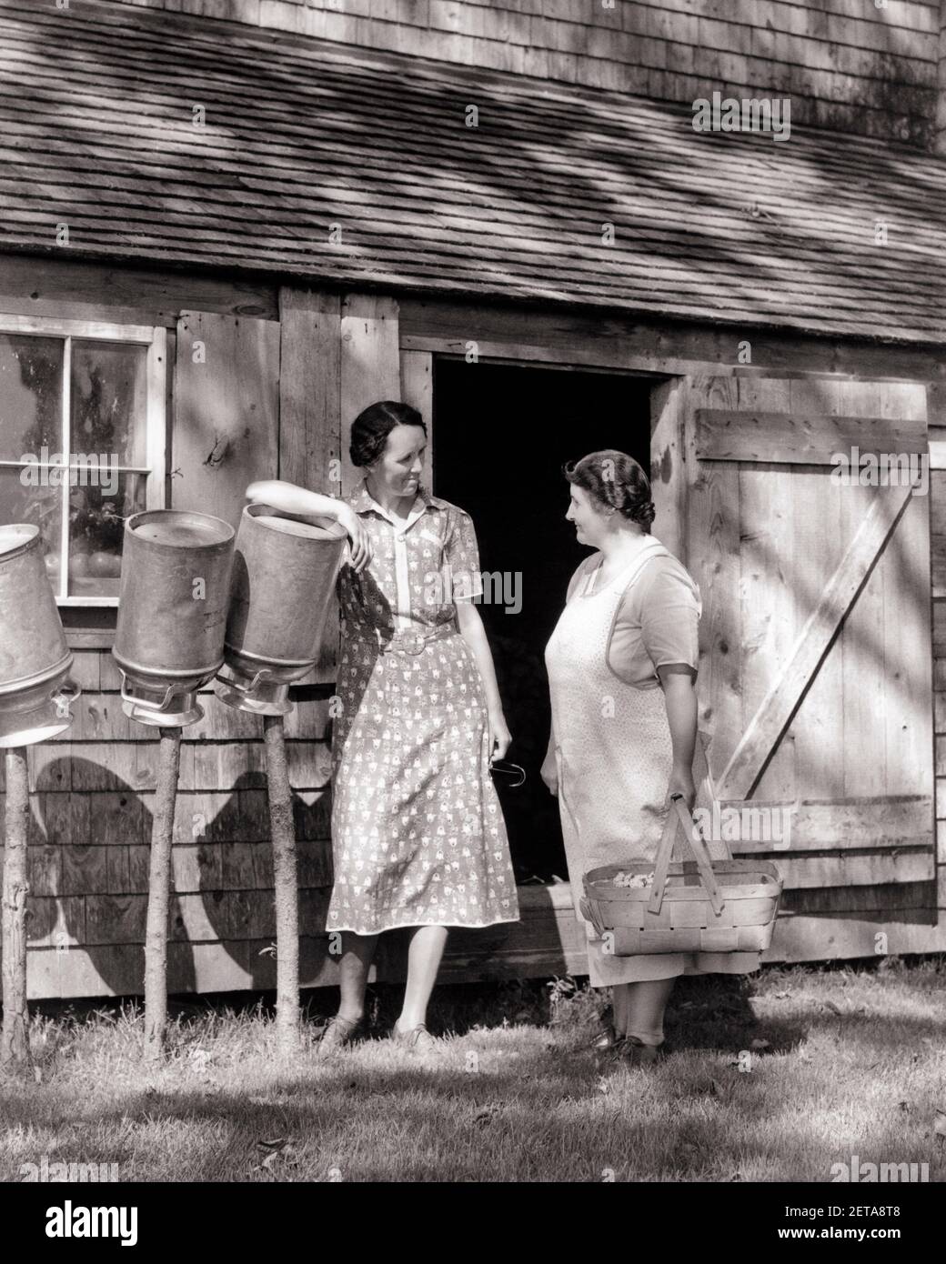 1930S ZWEI FRAUEN VERWICKELT IN GESPRÄCH NEBEN BAUERNHOF GEBÄUDE STEHEN STÖCKE MIT AUF DEN KOPF TROCKNENDEN METALLMILCHDOSEN KLATSCHEN - D4535 HAR001 HARS HOME LEBEN KOPIEREN RAUM FREUNDSCHAFT IN VOLLER LÄNGE DAMEN PERSONEN DOSEN LANDWIRTSCHAFT KOPF GESCHWISTER SCHWESTERN KLATSCH LANDWIRTSCHAFT B&W KLEIDER BUZZ GLÜCK TROCKNEN BAUERN BUSYBODY ERZÄHLEN ENGAGIERTE GESCHWISTER GESCHICHTEN VERBINDUNG NEBEN FREUNDLICHER TATTLER HÖRENSAGEN KOOPERATION MID-ADULT MID-ADULT FRAU PEGS GERÜCHTE ZWEISAMKEIT SCHWARZ UND WEISS KAUKASISCHE ETHNIE HAR001 ALTMODISCH Stockfoto