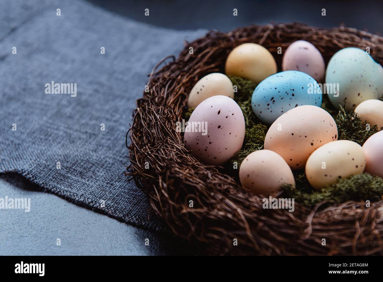 Nahaufnahme Weidennest mit Moos und farbigen Ostereiern auf grauer Leinenstoff-Serviette auf dunklem Hintergrund. Dunkle Stimmung. Frohe Ostern Stockfoto