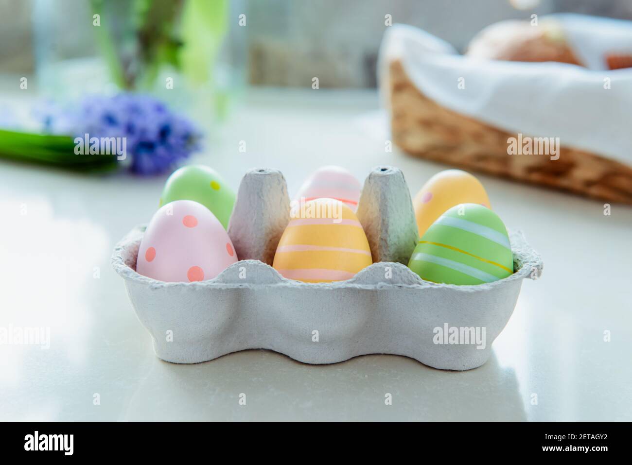Bunt bemalte Eier in einer Pappschachtel auf dem Hintergrund Aus frischer Hyazinthe Blume und Strohkorb mit festlichem Gebäck Auf weißem Küchentisch Stockfoto