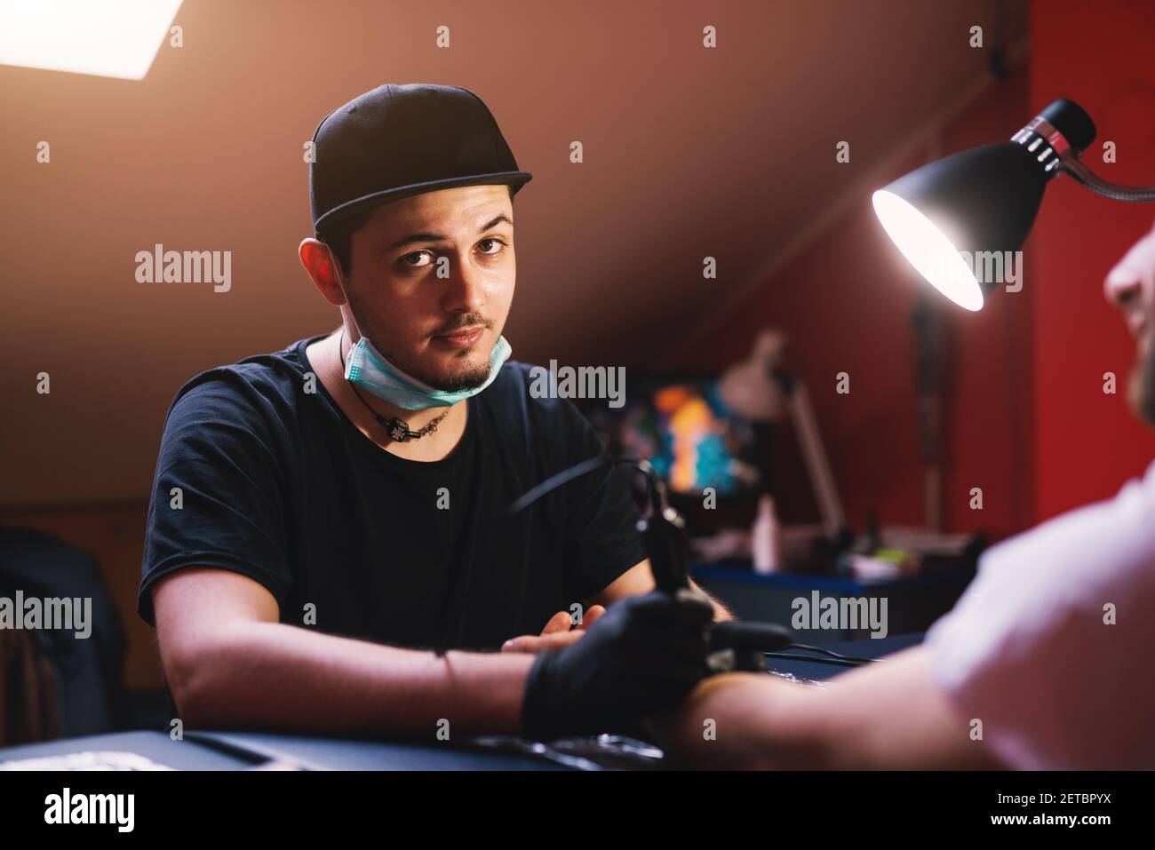 Profil des jungen neugierigen Tätowierers mit Hut auf, der in seinem Salon eine Pause vom Tätowieren macht. Stockfoto