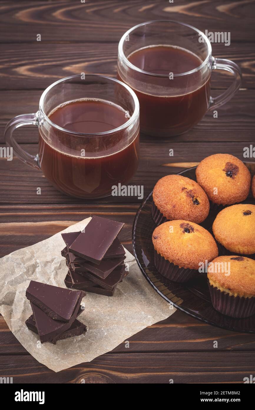 Stücke von dunkler Schokolade, Muffins und zwei Tassen mit heißem Kakao Getränk auf einem Hintergrund von braunen Holzbrettern. Das Konzept des romantischen Frühstücks. Getränke Stockfoto