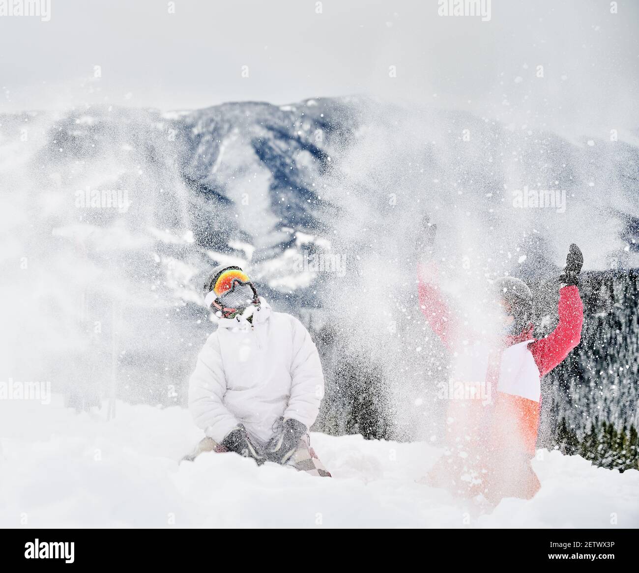 Junge Paar Skifahrer spielen im Schnee gegen faszinierende Landschaften im Hintergrund und werfen Schnee. Lebendige bunte Anzüge auf weißem Schnee. Konzept der Wintersport-Aktivitäten, Spaß und Beziehungen. Stockfoto
