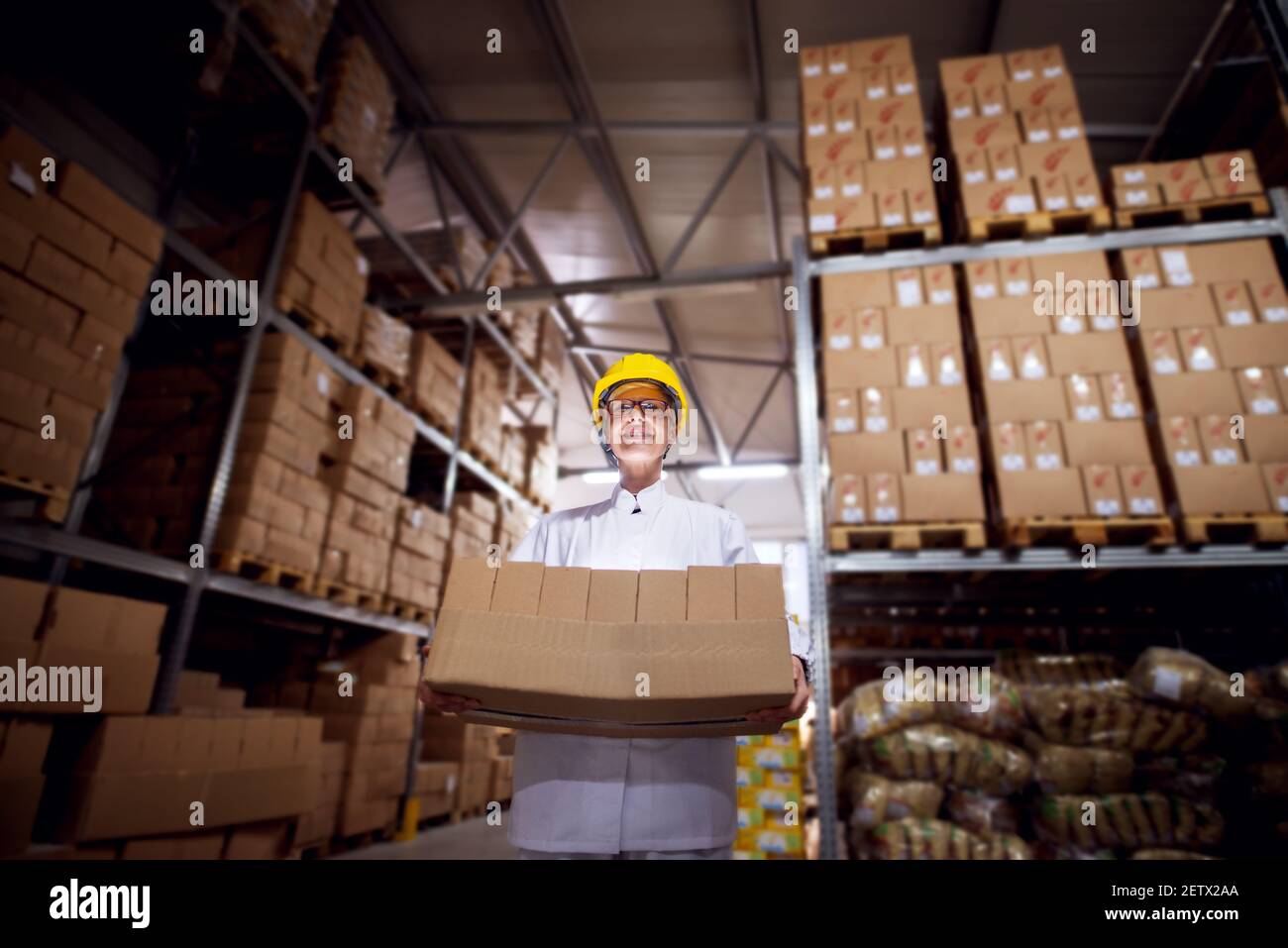 Junge angespannte Arbeiterin in sterilen Tüchern und gelbem Helm trägt einen sehr schweren Stapel brauner Pappkartons aus einem Lagerraum der Einrichtung. Stockfoto