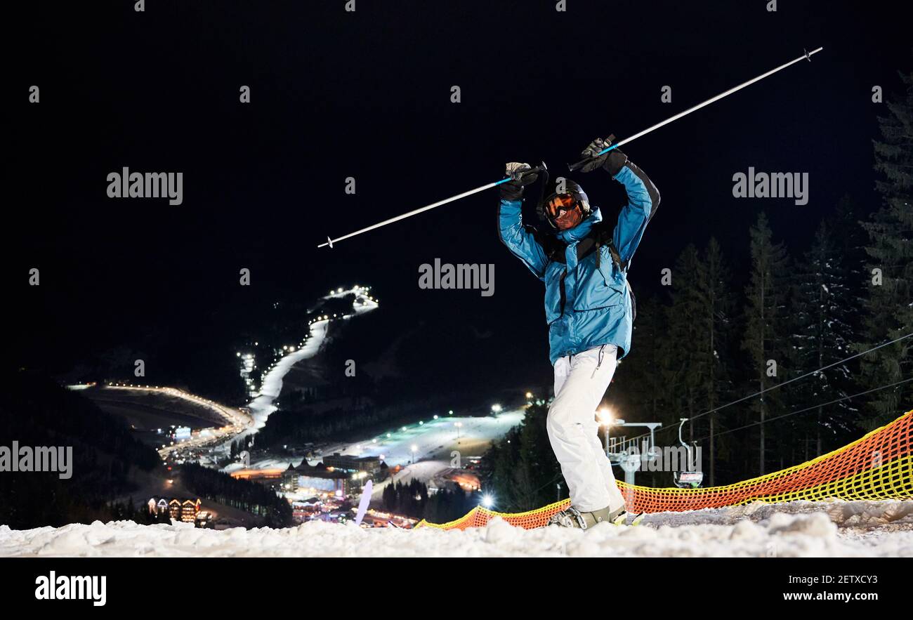 Der Skifahrer hält die Hände hoch und steht auf dem Hügel und bereitet sich auf eine Nachtfahrt vor. Schöne beleuchtete Skipisten sind unten auf Hintergrund in der Nacht. Konzept des Extremsports Stockfoto
