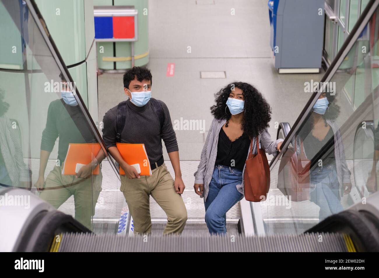 Ein junges lateinisches Paar, das eine Gesichtsmaske trägt, geht in die Rolltreppen des Bahnhofs oder der U-Bahn-Station. Neu normal bei öffentlichen Verkehrsmitteln. Stockfoto