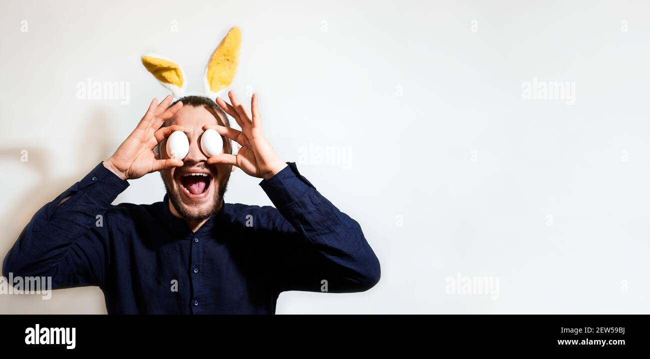 Charmanter kaukasischer Mann mit Bart mit gelben Kaninchenohren und zwei Hühnereiern für die Augen. Kreativer Osterhase auf weißem Hintergrund, Banner und viel Stockfoto