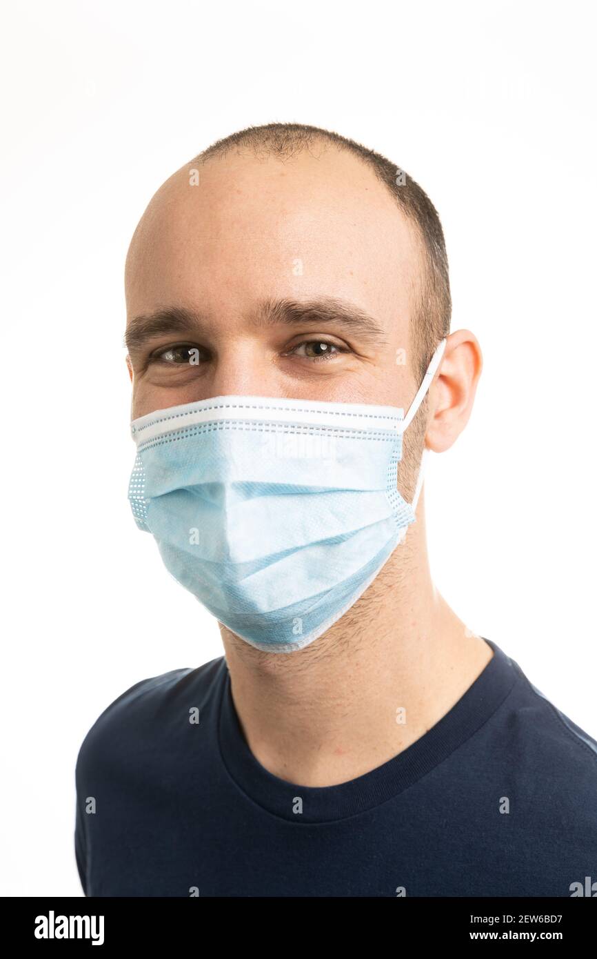Porträt eines kaukasischen Mannes, der ein blaues T-Shirt trägt und eine chirurgische Gesichtsmaske trägt, die mit den Augen lächelt. Weißer Hintergrund. Stockfoto