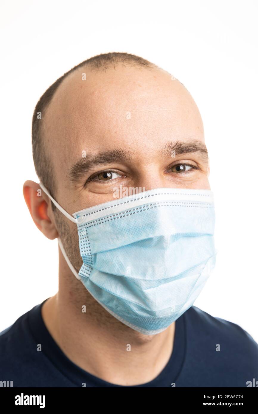 Porträt eines kaukasischen Mannes, der ein blaues T-Shirt trägt und eine chirurgische Gesichtsmaske trägt, die mit den Augen lächelt. Weißer Hintergrund. Stockfoto