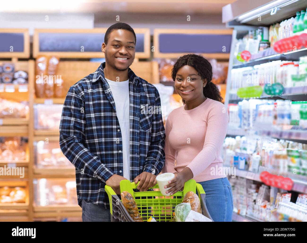 Portrait von positiven schwarzen Paar mit Trolley voller Produkte Blick auf die Kamera und lächelnd auf riesigen Supermarkt Stockfoto