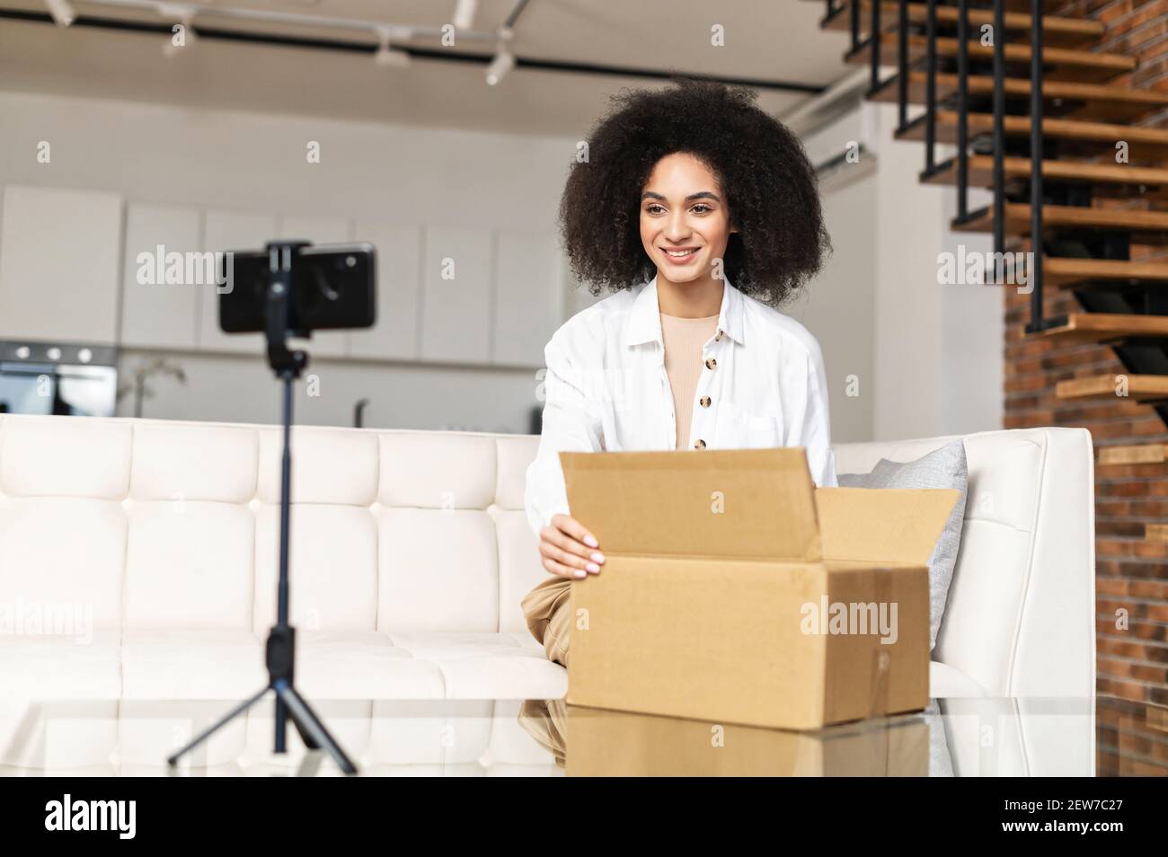 Zufrieden lächelnd junge afroamerikanische Bloggerin Frau auf Sofa sitzen Öffnen Paket Karton-Box, Auspacken empfangen Artikel aus Online-Shop, zeigt Auspacken Prozess für Anhänger in sozialen Medien Stockfoto