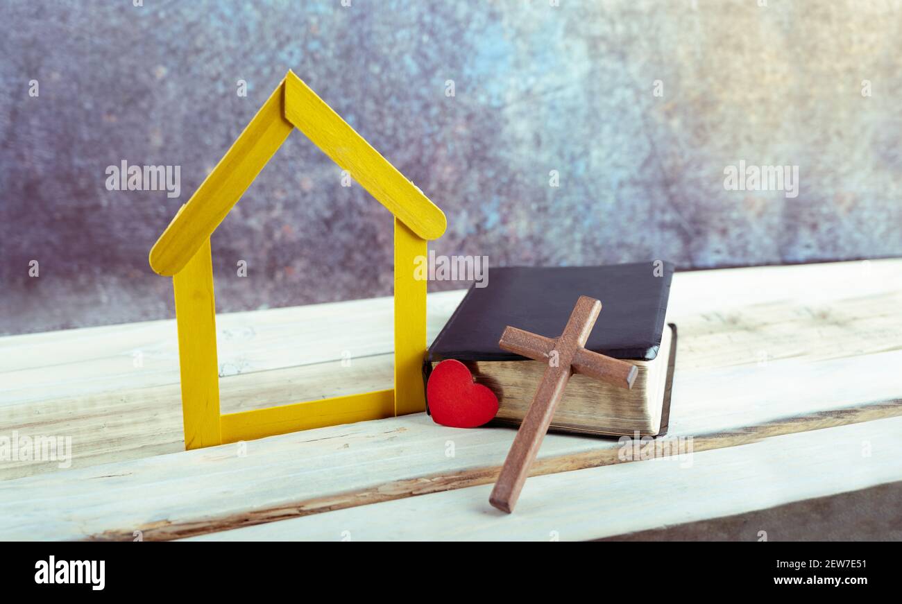 Herz und Holzkreuz lehnt an einer bibel, neben kleinen gelben Haus Holz auf weißem Hintergrund. Copy Space Stockfoto