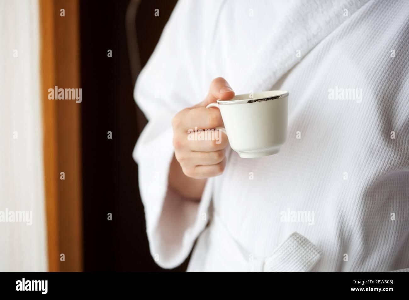 Ein Mann in einem weißen Frottee-Morgenmantel hält einen Kaffeebecher vor einem Fenster. Stockfoto