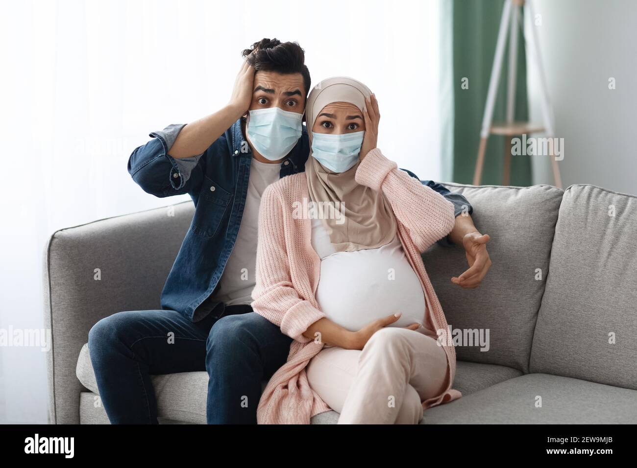 Schwangerschaft Während Der Pandemie. Schockiert Schwanger Muslim Paar In Masken Sitzen Auf Couch Stockfoto