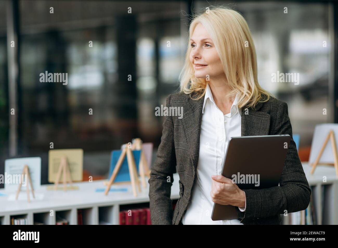 Stilvolle elegante reife Dame in formeller Kleidung stehen im Büro, Blick auf die Seite, denken über neue Projekt. Wunderschöne Geschäftsfrau arbeitet als Büroangestellter, hält Laptop in den Armen Stockfoto