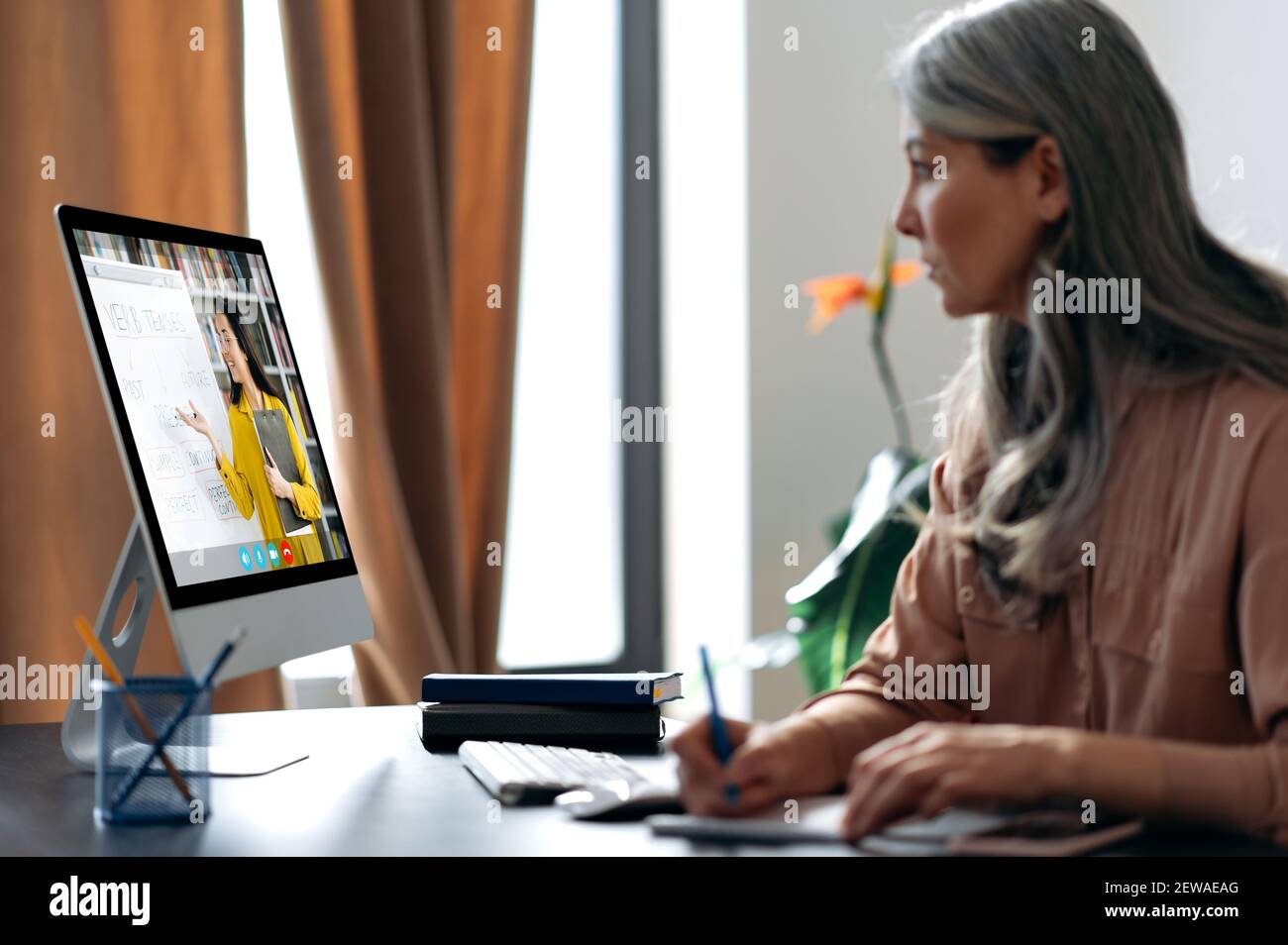 Fernunterricht. Konzentriert gealterte grauhaarige Frau, die online per Videoanruf studiert, nutzt App und Computer, sitzt an ihrem Schreibtisch, hört sich eine Online-Lektion an, auf dem Bildschirm ist der Lehrer in der Nähe Stockfoto