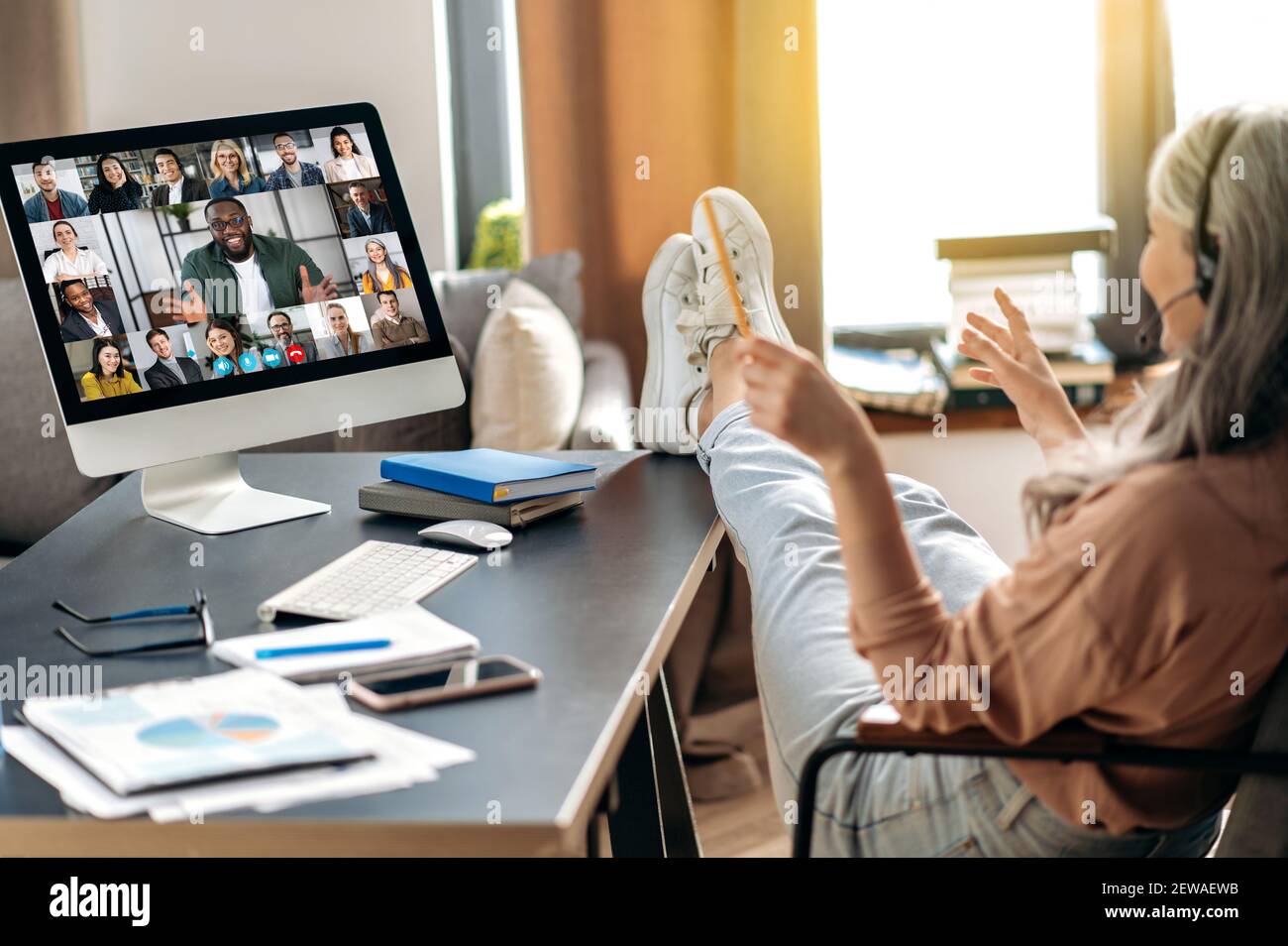 Moderne reife grauhaarige Frau kommunizieren mit Kollegen online per Videokonferenz verwendet Computer, sitzen in ihrem Home Office, werfen ihre Füße auf den Tisch, auf dem Bildschirm viele verschiedene Menschen Stockfoto