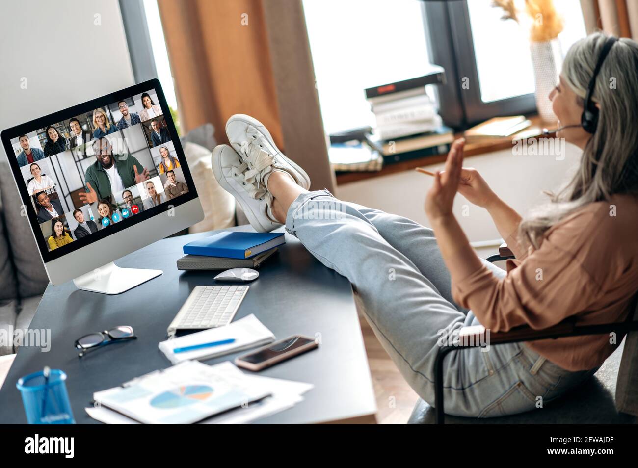 Online-Briefing. Moderne ältere grauhaarige Frau mit Kollegen online per Videoanruf kommunizieren nutzt App und Computer, sitzen in ihrem Home-Office, werfen ihre Füße auf den Tisch Stockfoto