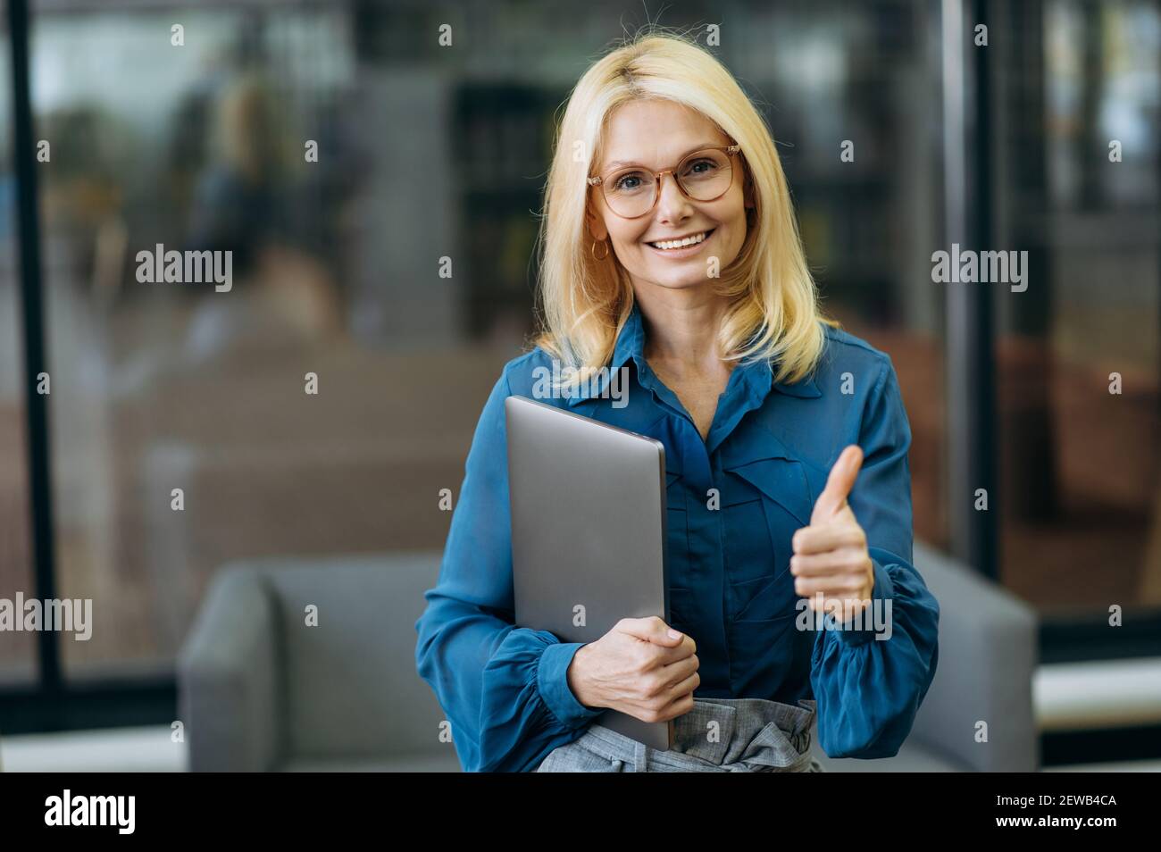 Porträt der glücklichen kaukasischen charmante Geschäftsfrau in Brillen am Arbeitsplatz, zeigt Daumen nach oben, hält einen Laptop, Blick auf die Kamera mit freundlichem Lächeln Stockfoto