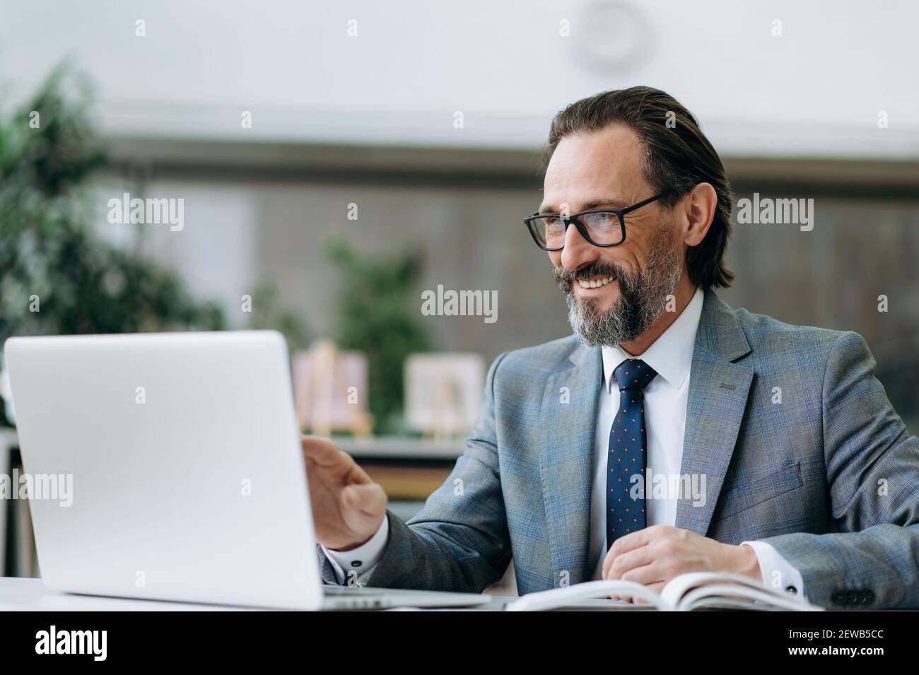 Fröhliche mittleren Alters Geschäftsmann ist mit Laptop, mit Online-Konferenz. Erfolgreicher männlicher Mitarbeiter, der per Videoanruf mit Kollegen spricht, Ideen diskutiert Stockfoto