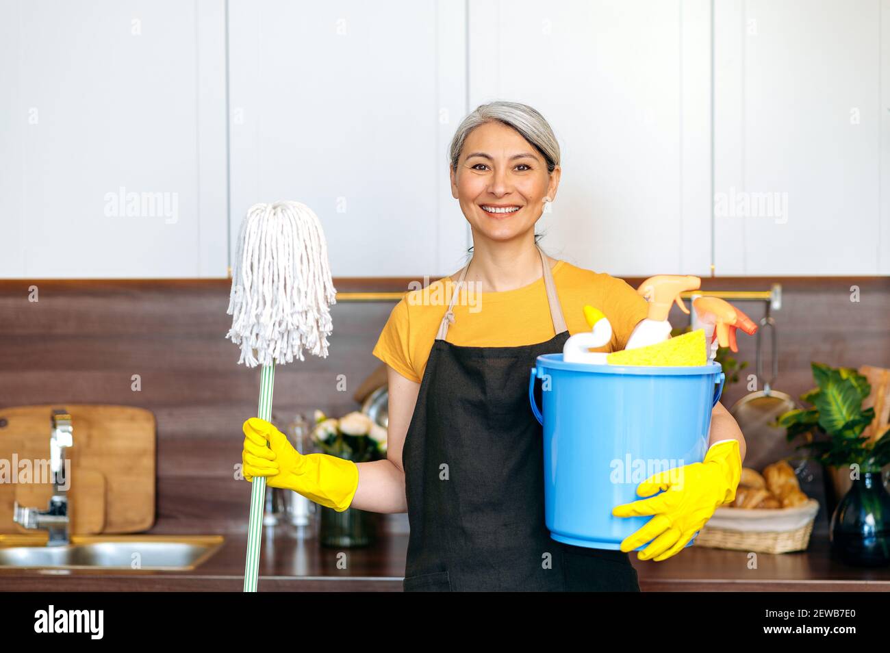 Reinigungsservice. Freundliche reife grauhaarige Putzfrau, hält einen Eimer mit Waschmitteln und Lumpen, Mopp in der anderen Hand, steht an der Küche, schaut auf die Kamera und lächelt, bereit zu reinigen Stockfoto