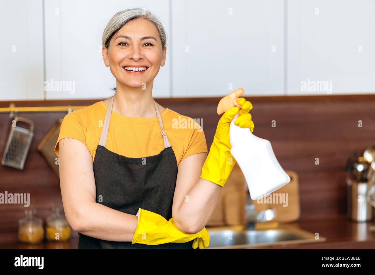 Zufrieden lächelnd reife asiatische Hausfrau oder Putzfrau in gelben Handschuhen und einer Schürze, hält Waschmittel, bereit zu Hause oder im Büro Reinigung, schaut auf die Kamera Stockfoto