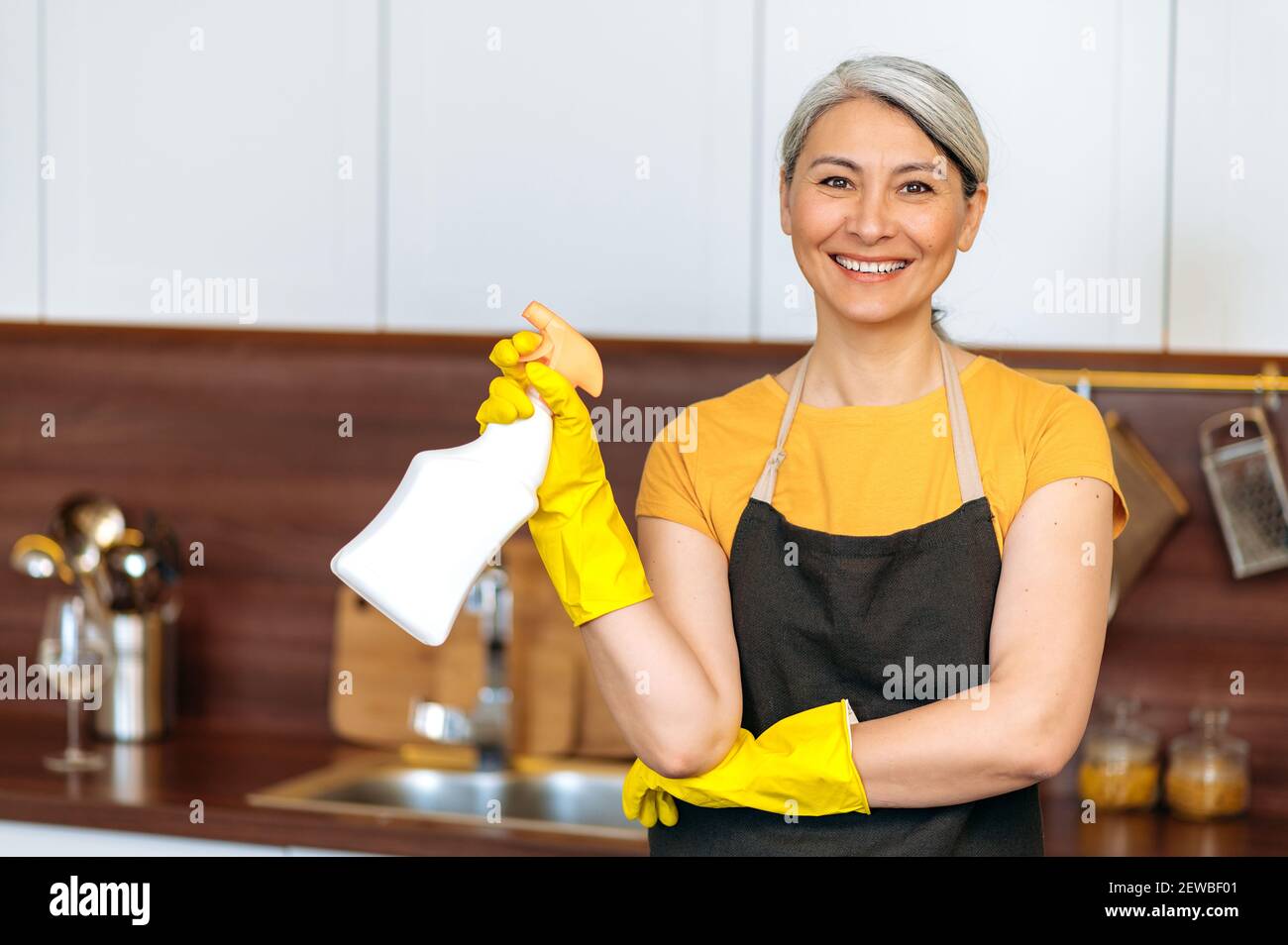 Zufrieden lächelnd reife asiatische Hausfrau oder Putzfrau in gelben Handschuhen und einer Schürze, hält Waschmittel, bereit zu Hause oder im Büro Reinigung, schaut auf die Kamera Stockfoto