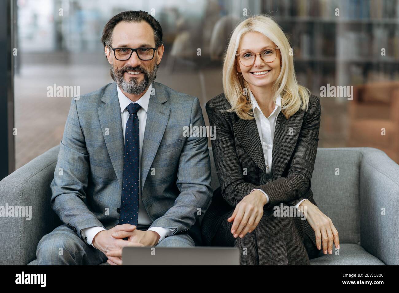 Porträt einer erfolgreichen männlichen und weiblichen Angestellten, die auf der Couch sitzen, lächelnd auf die Kamera schauen. Geschäftspartner mittleren Alters tragen stilvolle Anzüge und arbeiten im Büro zusammen Stockfoto