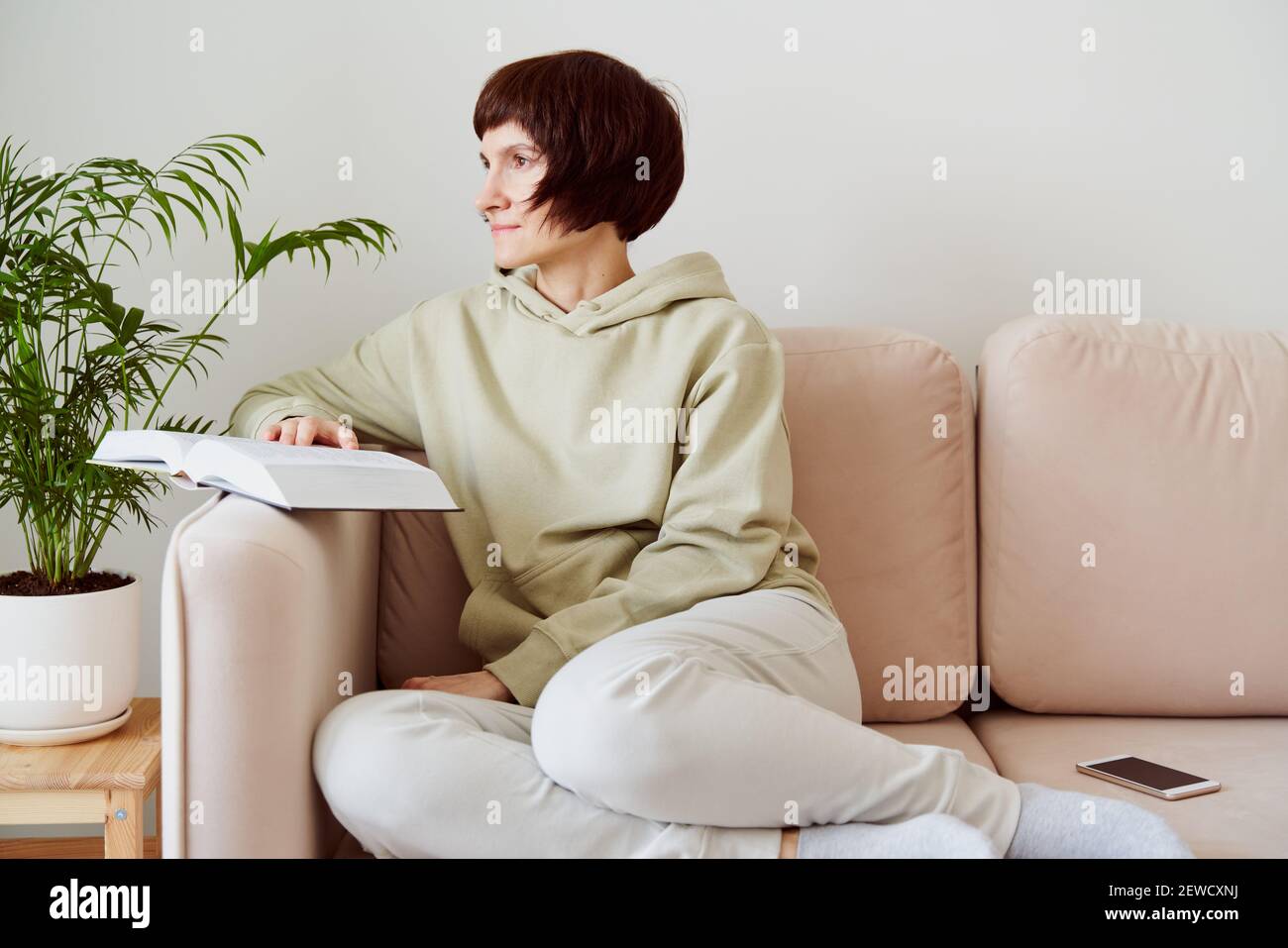 Reife Frau, die Buch liest, auf dem Sofa sitzend, zum Fenster schauend, träumend und denkend. Digitale Entgiftung Stockfoto