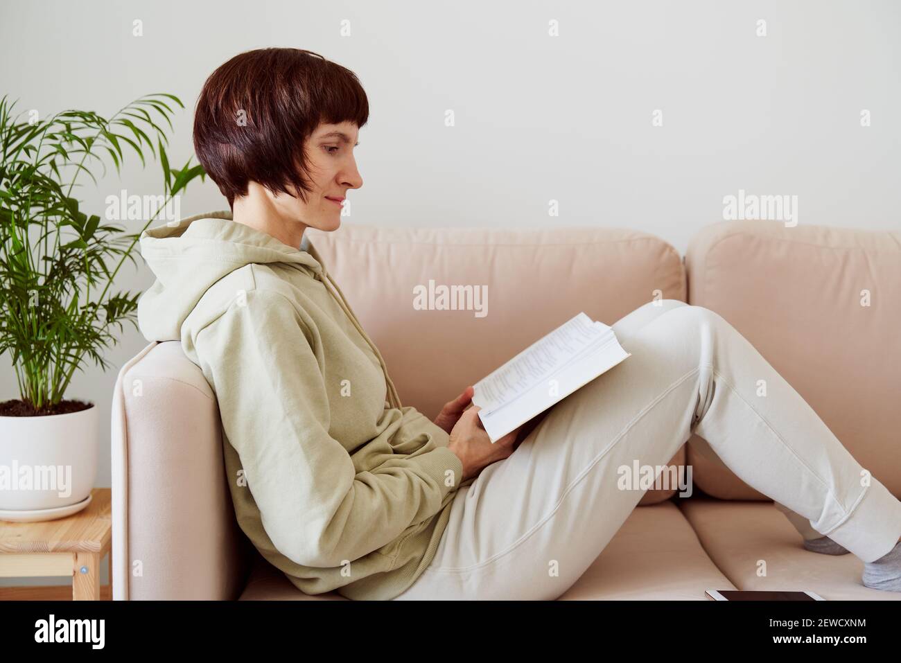 Reife Frau liest Buch sitzend auf Sofa, allein denkend. Digitales Detox-Konzept, Freizeit für sich Stockfoto