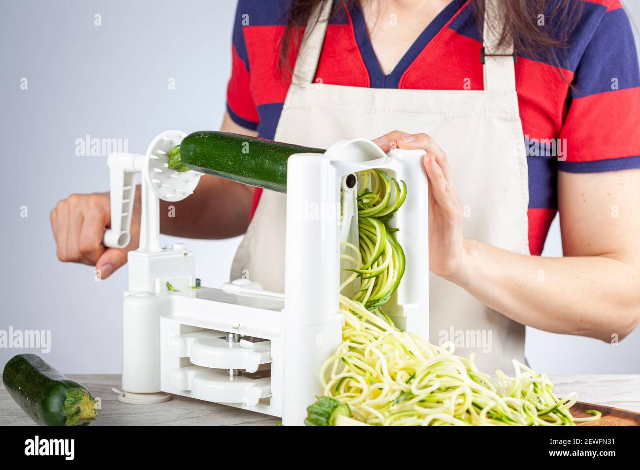 Eine kaukasische Frau trägt Schürze dreht den Griff eines Gemüse Spiralizer, Slicer, um hausgemachte Zucchini-Nudeln auf Küche Arbeitsplatte zu machen. Dies Stockfoto