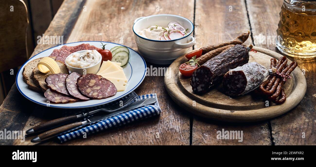 Appetitlich geräucherte Wildwurst und Käsescheiben Holztisch mit Besteck und Becher Bier Stockfoto